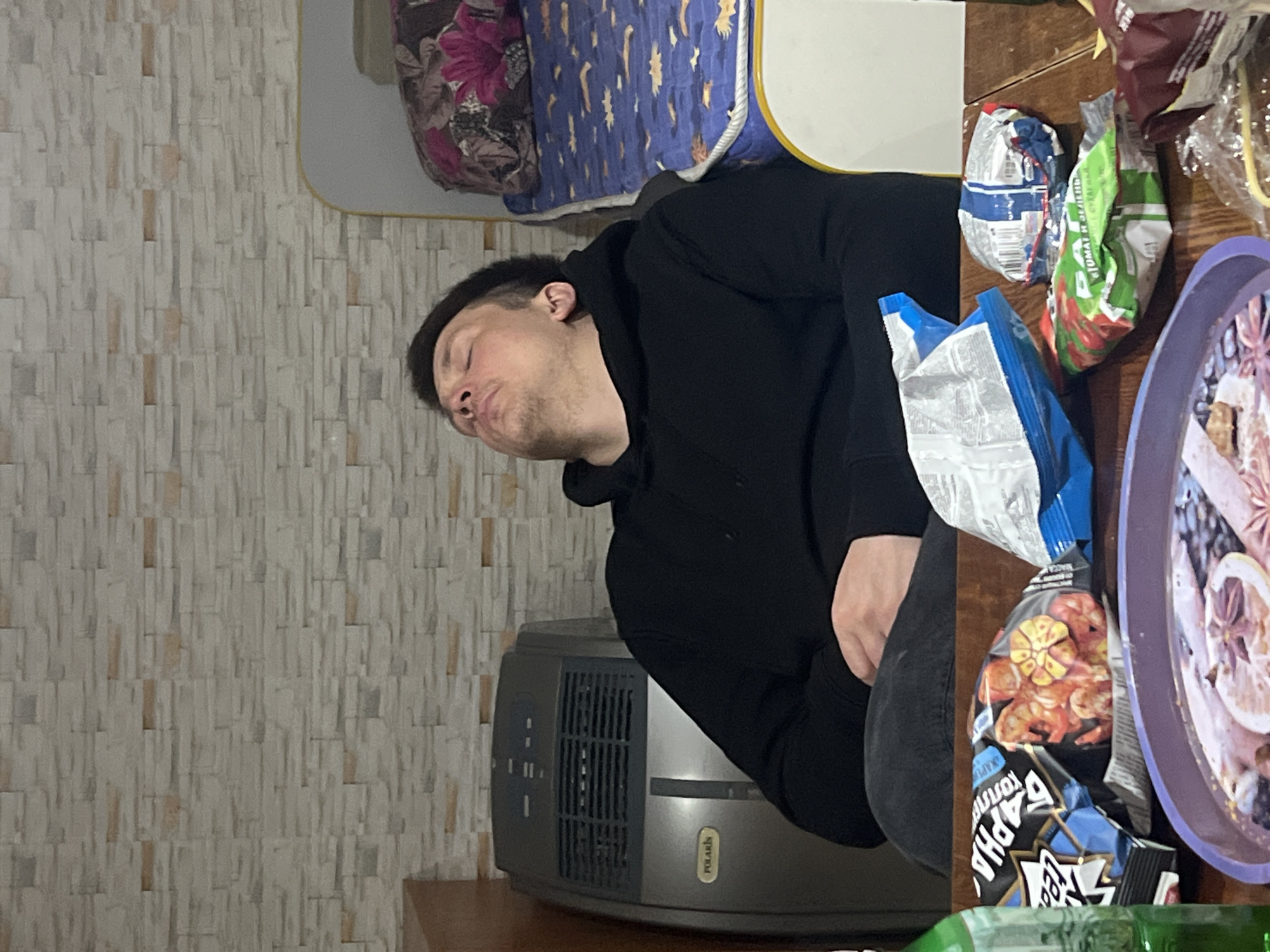 A man in a black hooded sweatshirt is asleep on a table, resting his head near a bed. On the table are various snack items, including multiple bags of chips and a platter with cut fruit. A portable heater with the brand name "Polaris" is positioned near the man. The scene is indoors, likely in a home setting. The wall has a patterned tile appearance. The location is Chaikovsky, Russia.