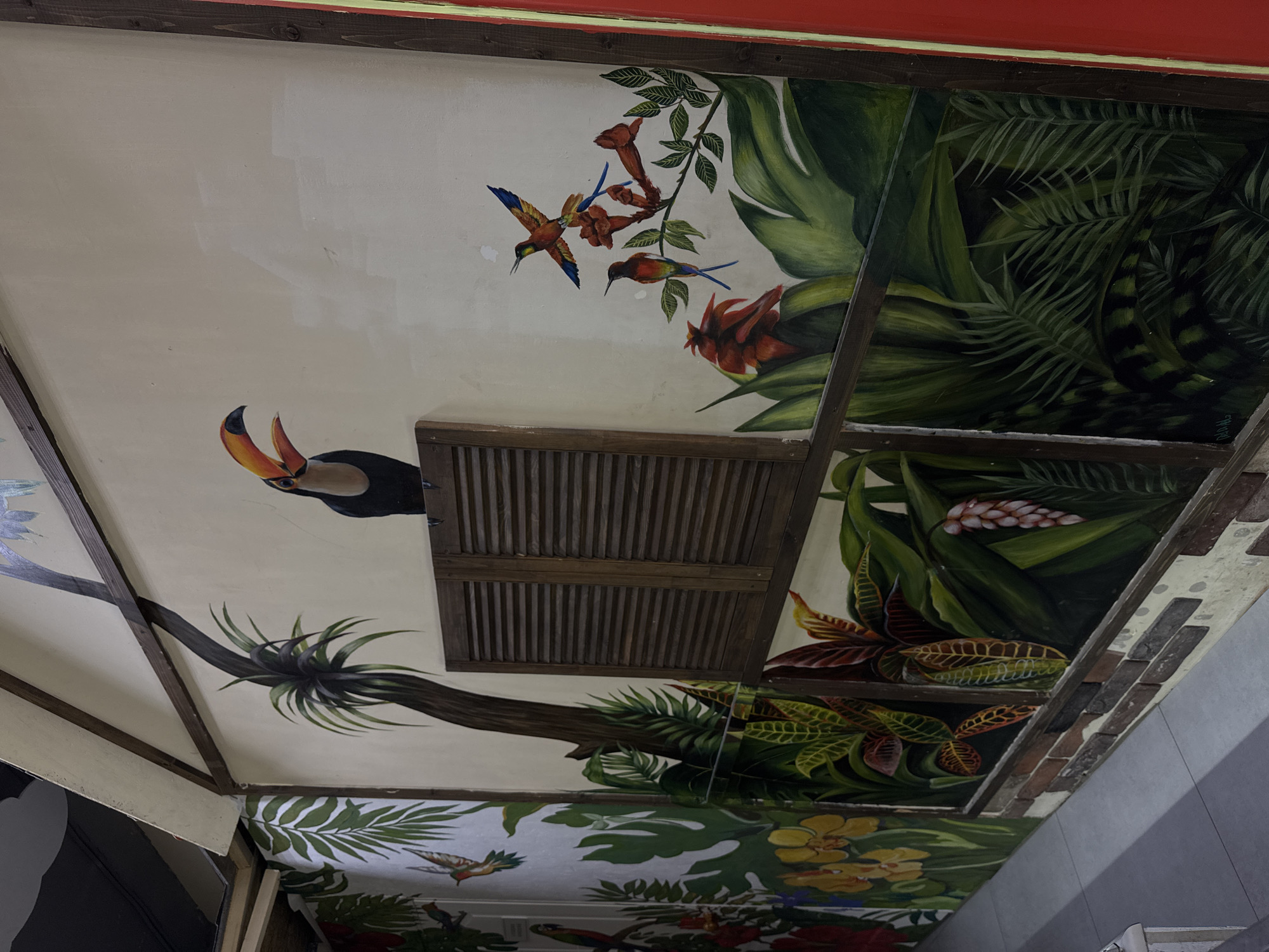 The image shows a mural painted on a wall, depicting a tropical jungle scene. The mural features various elements such as a toucan perched on a branch, hummingbirds flying amongst flowers, and lush green foliage. There are also wooden shutters integrated into the mural, resembling a window. The style of the mural is realistic and colorful, creating a vibrant and immersive atmosphere. The environment appears to be indoors, possibly a restaurant or a themed establishment, given the presence of a walkway or corridor in the foreground. The overall impression is that of a tropical oasis brought to life on a wall.