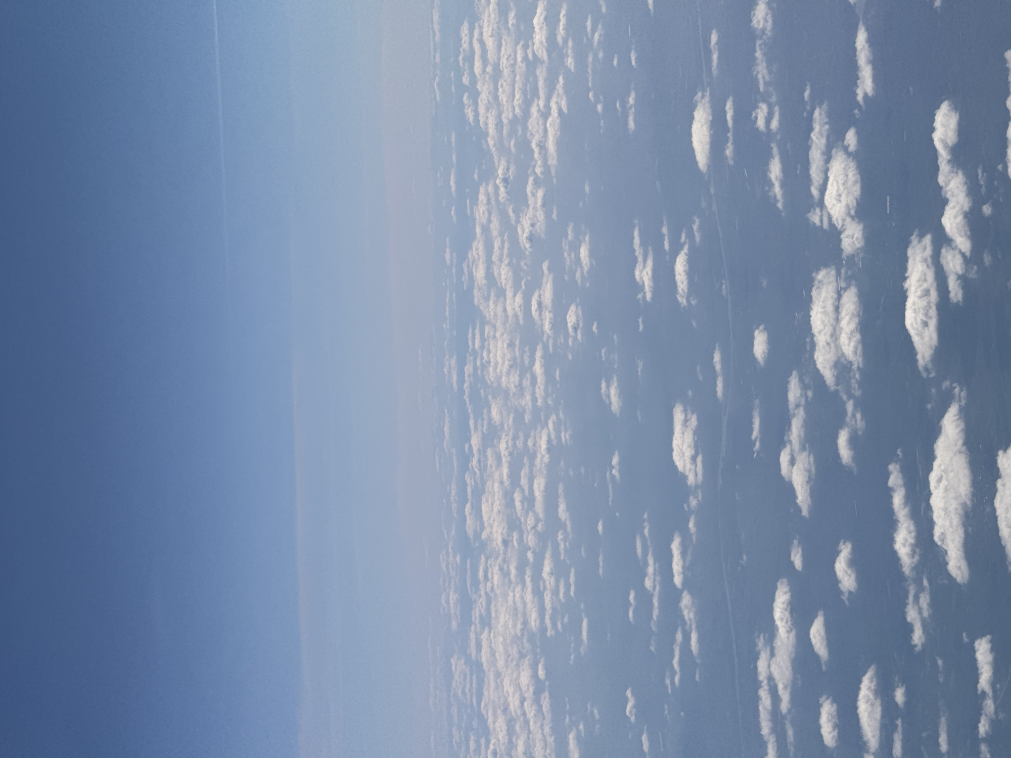 This media file presents an aerial view of the sky and clouds, likely captured from an aircraft. The image is composed of two main sections: a clear blue sky on the left and a field of scattered white clouds on the right.
The left half of the image features a bright, deep blue sky, exhibiting a subtle gradient that lightens towards the center. A faint, thin white contrail (vapor trail) from an aircraft is visible as a straight line stretching horizontally across the upper-left quadrant of this clear sky area.
The right half of the image is filled with numerous small, fluffy white cumulus clouds. These clouds are scattered across a lighter blue background, forming an expansive, textured layer. A very subtle, faint linear mark, possibly a distant atmospheric feature or another contrail, can be seen running diagonally through the cloud field.
The scene depicts a daytime setting, characterized by ample natural light and vibrant blue and white colors. The weather appears clear with scattered cloud cover. There are no visible people, objects besides the clouds and contrail, or any discernible text within the image. The perspective offers a sense of vastness and serenity. While the context mentions Sofia, Bulgaria, no specific geographical cues are visible to confirm this location.