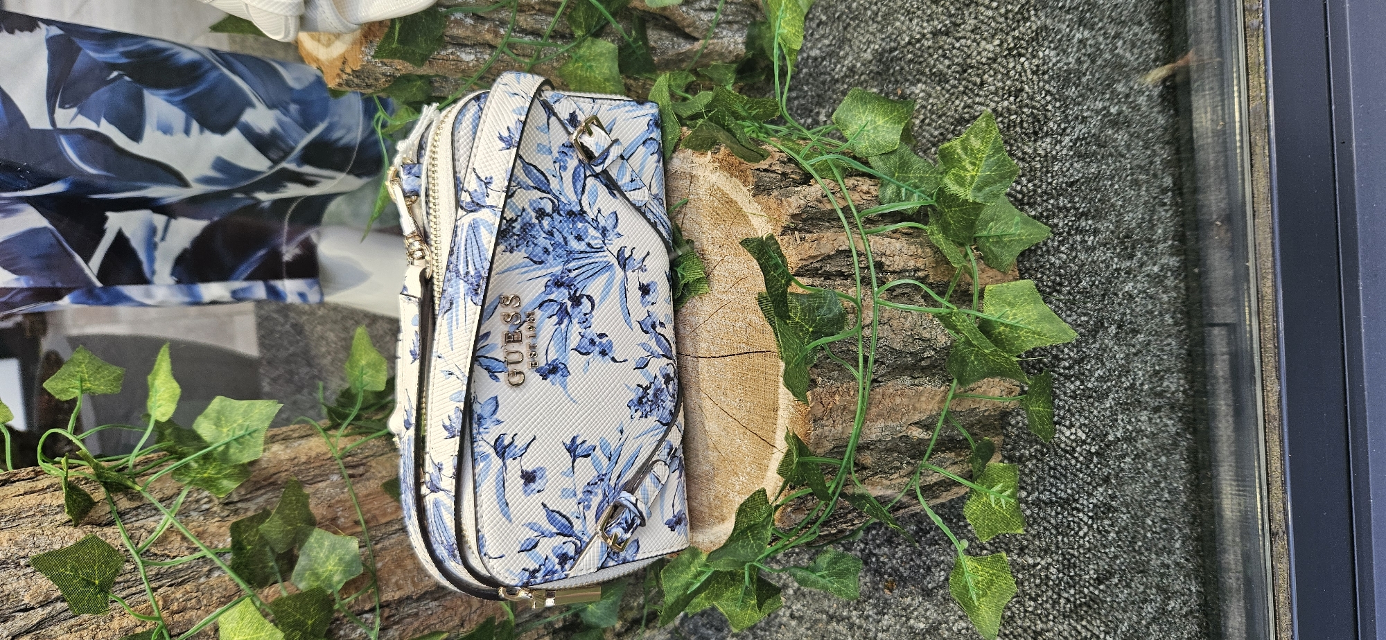 A white and blue floral-patterned Guess wallet is displayed on a rustic wooden log. Green artificial ivy vines are draped around the log and wallet. In the background, a blurred image of a fabric with a blue and white leaf pattern is visible, along with a gray carpeted surface. The scene appears to be an indoor display, possibly in a store or window, given the presence of a reflective surface and a dark frame on the right side. There are no people visible. The visible text on the wallet reads "GUESS EST 1981". The lighting suggests it is daytime.