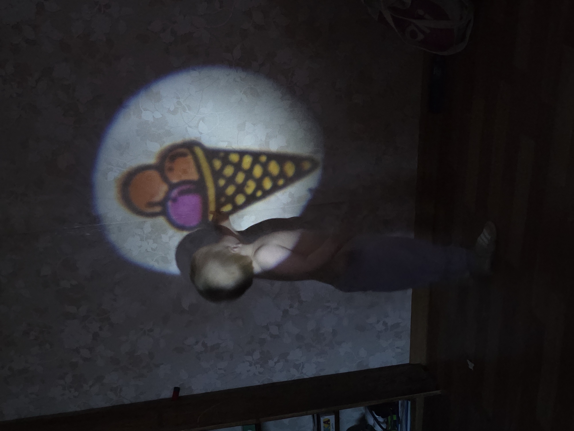 An interior room in a residence within the city of Chaikovsky, Russia, is depicted under dim lighting, with a single bright, circular projection on a light-colored, patterned wallpapered wall. A young child with light-colored hair, appearing bare-chested and wearing light-colored pants, stands facing the wall and looking upwards. The child's upper body and head are within the illuminated circle, creating a partial shadow that interacts with the projected image. The projection displays a cartoon-style image of an ice cream cone containing three scoops: two brown/orange and one purple, all within a yellow cone patterned with brown dots. To the immediate left of the child, a dark wooden bookshelf is visible, containing various items including what appear to be books or toy boxes with colorful labels. The floor is dark wood. In the lower right corner, a blurry, light-colored bag with red accents is partially visible on the floor.
