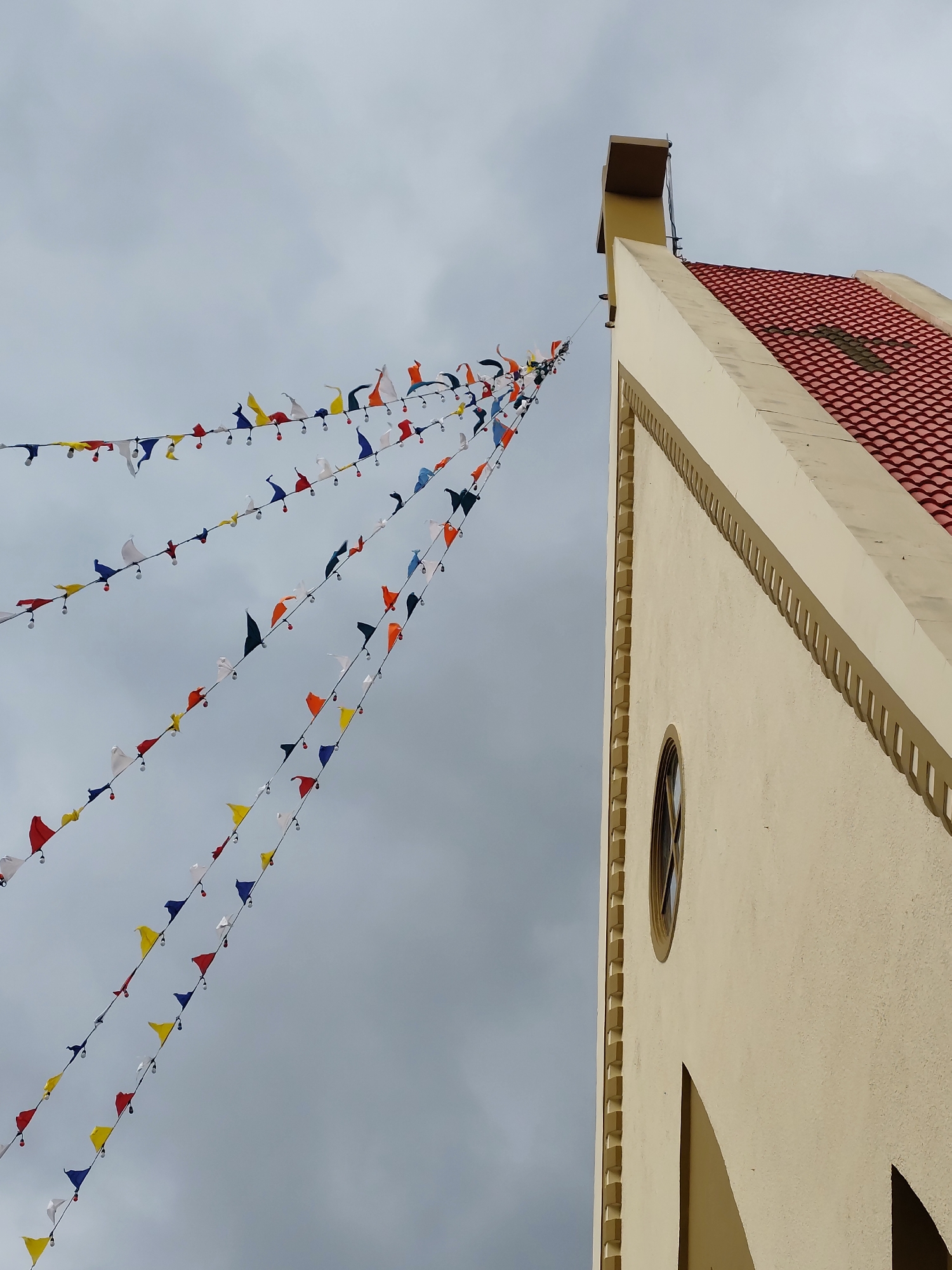 A low-angle shot shows a building and strings of flags under an overcast sky. The building is beige with a red tile roof and a circular window. Strings of colorful pennant flags are strung from the building's top. The sky is grey. The scene appears to be in Da Lat, Vietnam, known for its French colonial architecture and colorful decorations. The flags suggest a festive occasion or celebration. No people are visible.