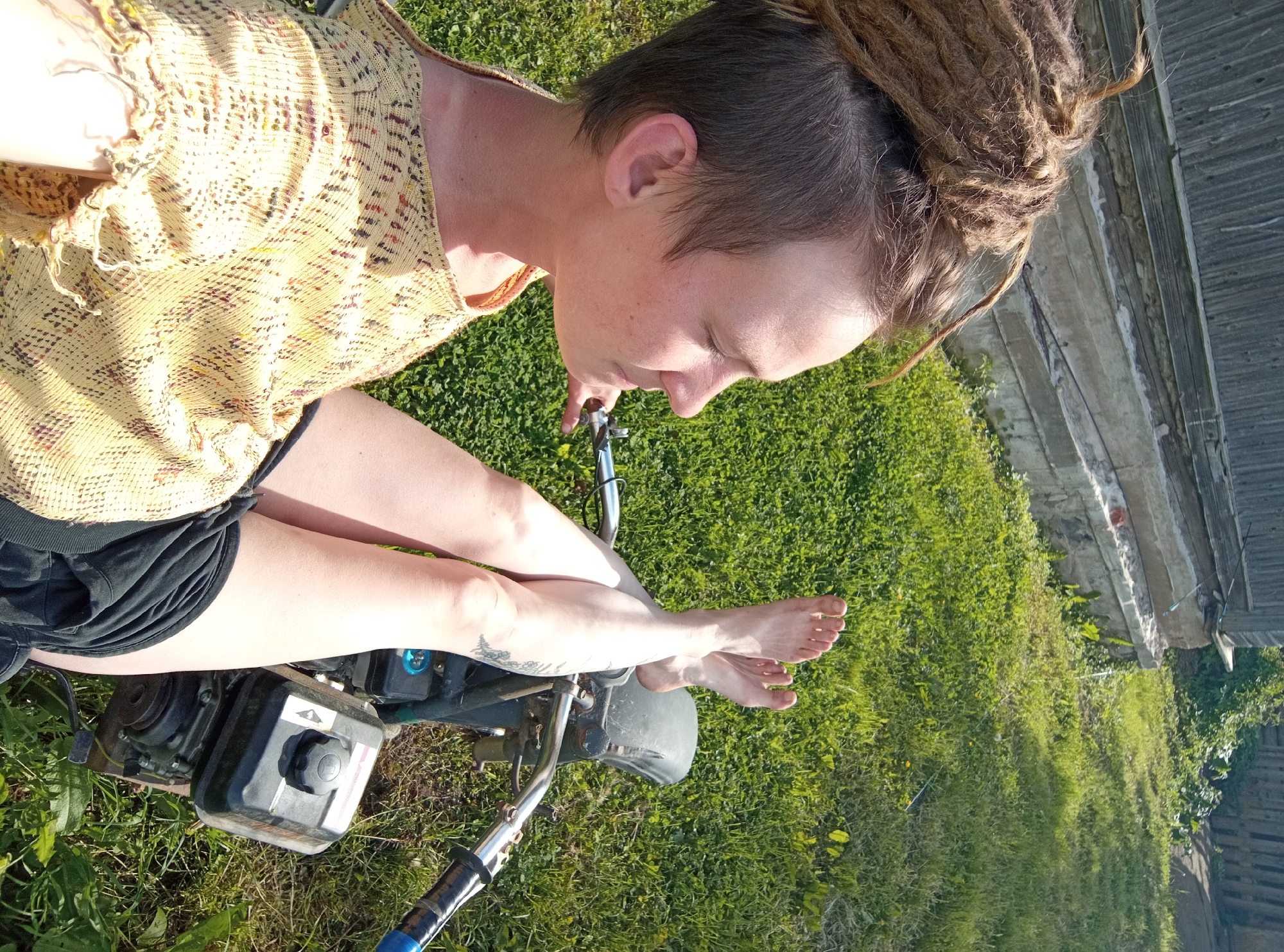 An overhead, slightly angled shot captures a person sitting barefoot on a small motorized vehicle, likely a mini-bike or scooter, in a grassy outdoor setting. The person, an adult with dreadlocks pulled back and shaved sides, wears a light-colored, patterned sleeveless top and dark shorts. Their legs are crossed at the ankles, and a small tree tattoo is visible on their left lower leg. They are looking down towards the engine of the vehicle, which features a black fuel cap with a white arrow symbol indicating direction or function.
The scene is brightly lit by strong sunlight, casting shadows on the green grass and the person's clothing, suggesting a sunny day. In the background, a rustic wooden building or fence is partially visible, hinting at a rural or semi-rural environment. Lush green foliage surrounds the structure. The person appears to be relaxed or observing something on the vehicle. The overall impression is one of a calm, sunny moment outdoors in Ostrovtsy, Russia.