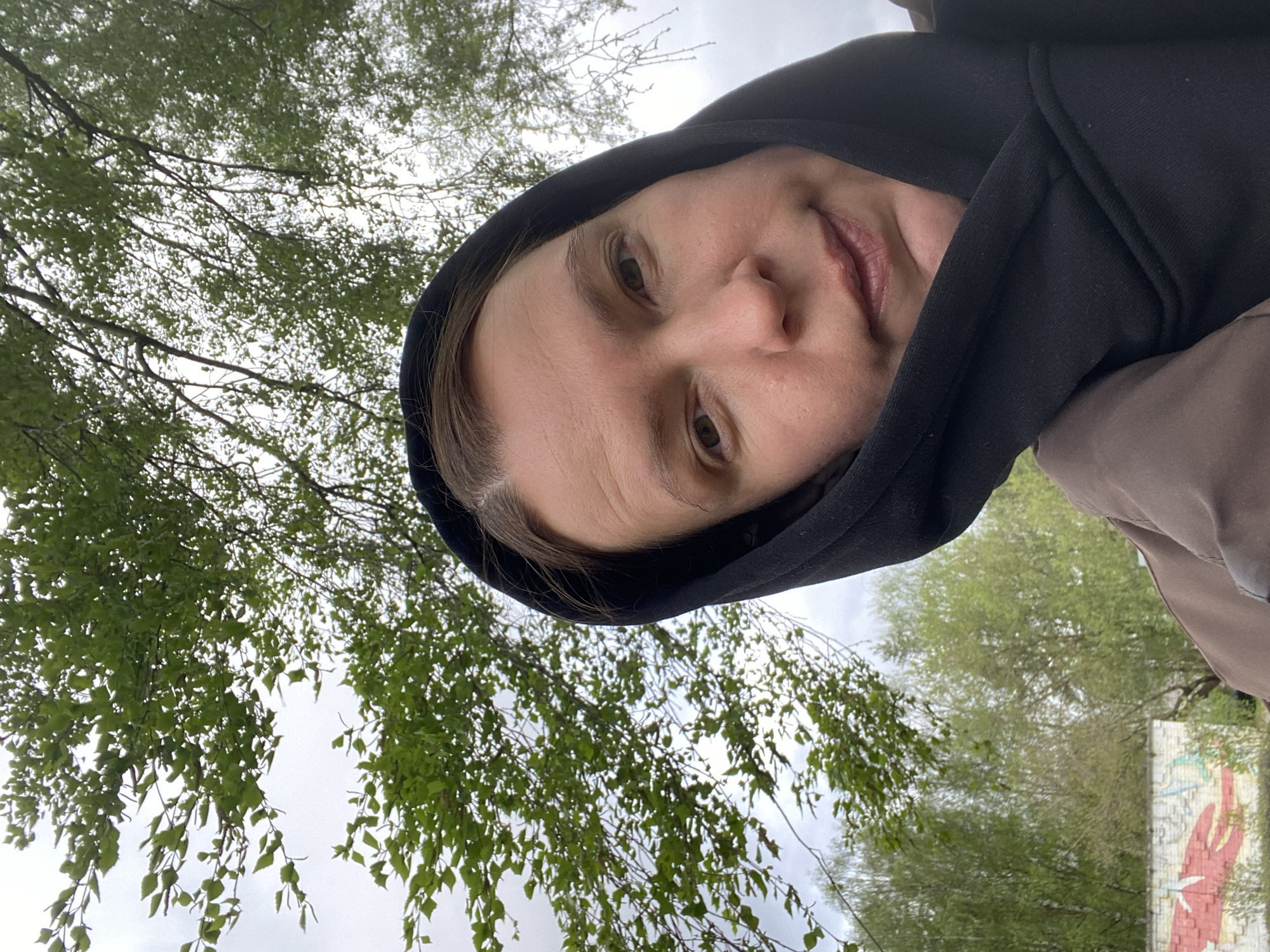 A woman in a black hooded jacket is angled toward the camera, with trees in the background. The location appears to be the city of Chaikovsky, Russia, as suggested by the presence of a large, painted mural of a red lobster on a textured background of brick. The scene is outdoors under cloudy skies, with the woman's face and the foliage of the trees being the most prominent elements.
