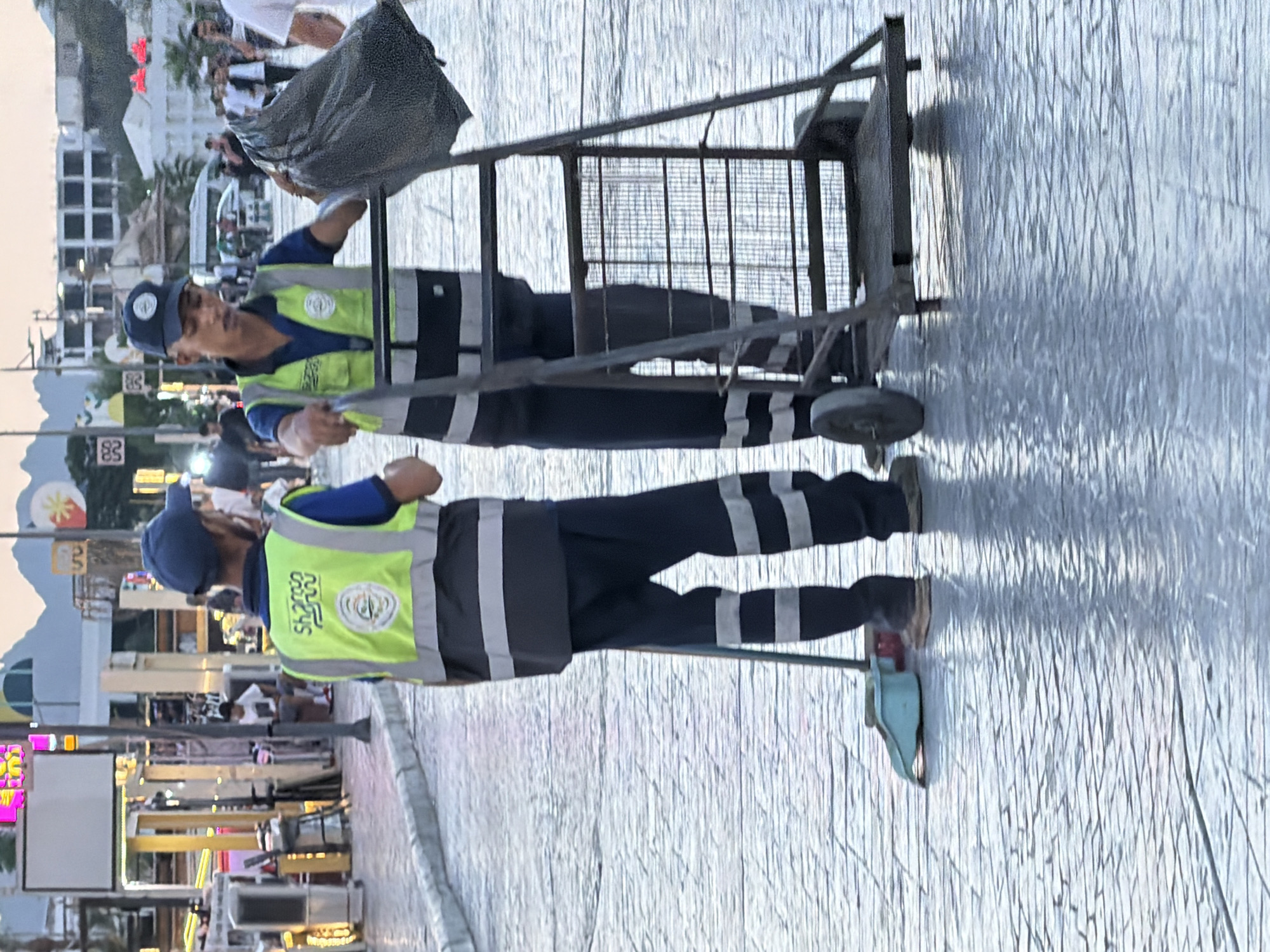 The image depicts two sanitation workers in Sharm El Sheikh, Egypt, on a paved pedestrian area. Both individuals wear high-visibility vests over blue shirts and dark pants with reflective stripes. The worker on the left holds a broom and dustpan, suggesting an ongoing cleaning task. The worker on the right is next to a trolley and is holding a large black trash bag, implying waste collection. In the background, buildings, streetlights, and other pedestrians are visible, indicating an urban environment, likely a commercial or tourist area. The ground appears wet, possibly from recent cleaning or rain, reflecting the ambient light. The overall scene captures a moment of daily municipal work in a lively city setting.