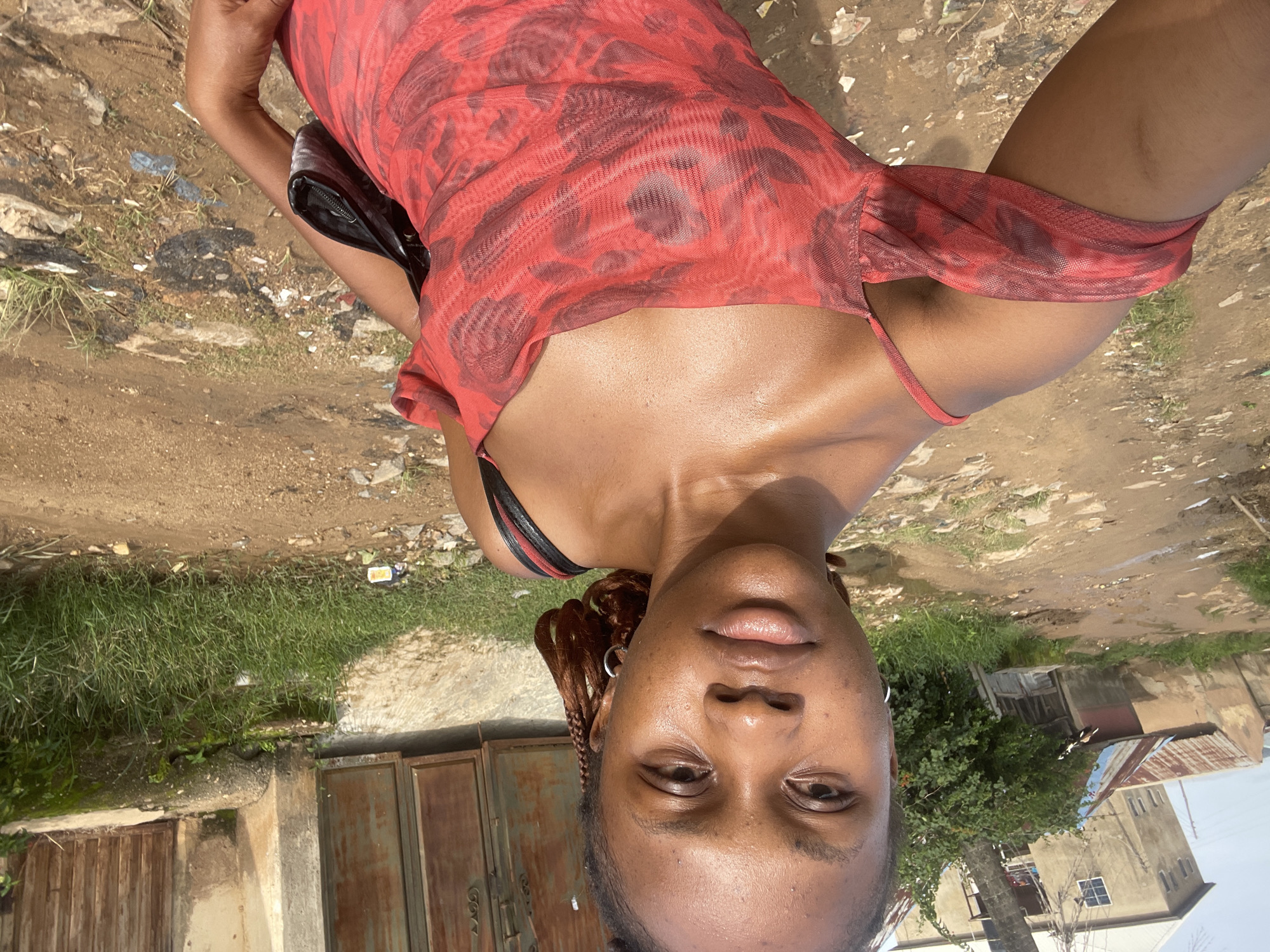 The media file shows a close-up, overhead shot of a Black woman posing outdoors. She is wearing a red and black patterned dress and a black bra. Her hair is braided and she is wearing earrings. The background consists of a dirt path, tall grass, and some buildings, suggesting a rural or semi-urban setting. The lighting indicates it is daytime, with the sun casting shadows. The woman appears relaxed and is looking towards the camera with a slight smile. The scene seems to be in Rafin Par, Nigeria, as indicated by the location context. There is no visible text in the image.
