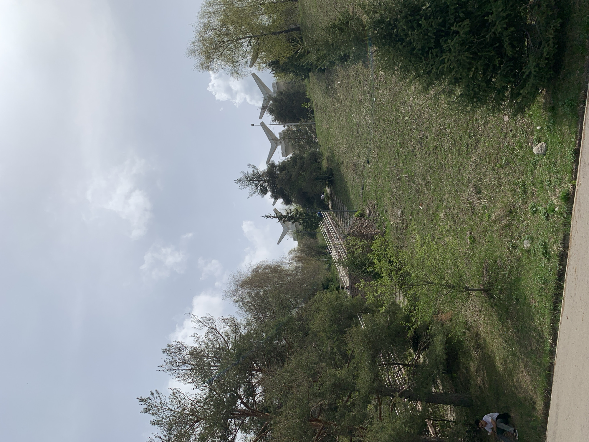 The image shows a hillside in Almaty, Kazakhstan, with the "Three Heroes" monument visible in the background. The monument consists of three large concrete structures resembling stylized wings or blades. The hillside is covered in grass and trees. A metal railing runs along a path or steps leading up the hill. In the foreground, at the bottom of the frame, are two people, one wearing a white shirt and dark pants, the other wearing a white shirt and light pants. A vertical concrete structure is along the side. The sky is overcast with white clouds.