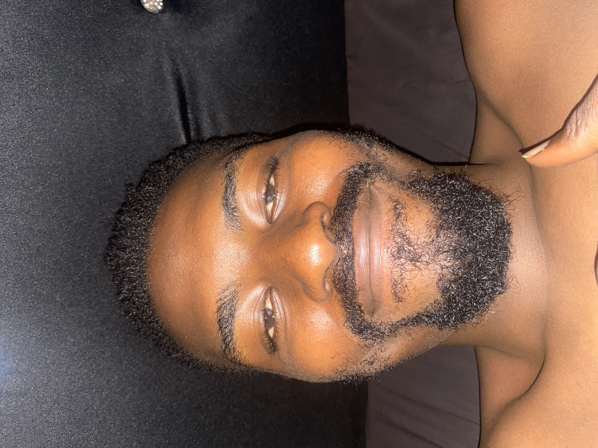 A close-up, rotated portrait shows an adult individual with dark skin, likely photographed in Barakin Akawo, Nigeria. The person is lying down, facing upwards and looking directly at the camera, with their head positioned towards the left side of the frame. They have short, curly dark hair and a full, short beard. Reflections on their skin, particularly across the forehead and nose, suggest an artificial light source. The eyes are open, conveying a calm or neutral expression.
The background is dark, appearing to be a soft, black fabric, possibly part of a bed or pillow. A small, shiny, silver-colored stud is visible in the upper left corner of the background. On the right side of the frame, a bare shoulder and upper arm are partially visible, with a finger gently resting near the neck/shoulder area. The overall lighting is dim, indicating an indoor setting, possibly during the evening or night. There is no visible text in the image.