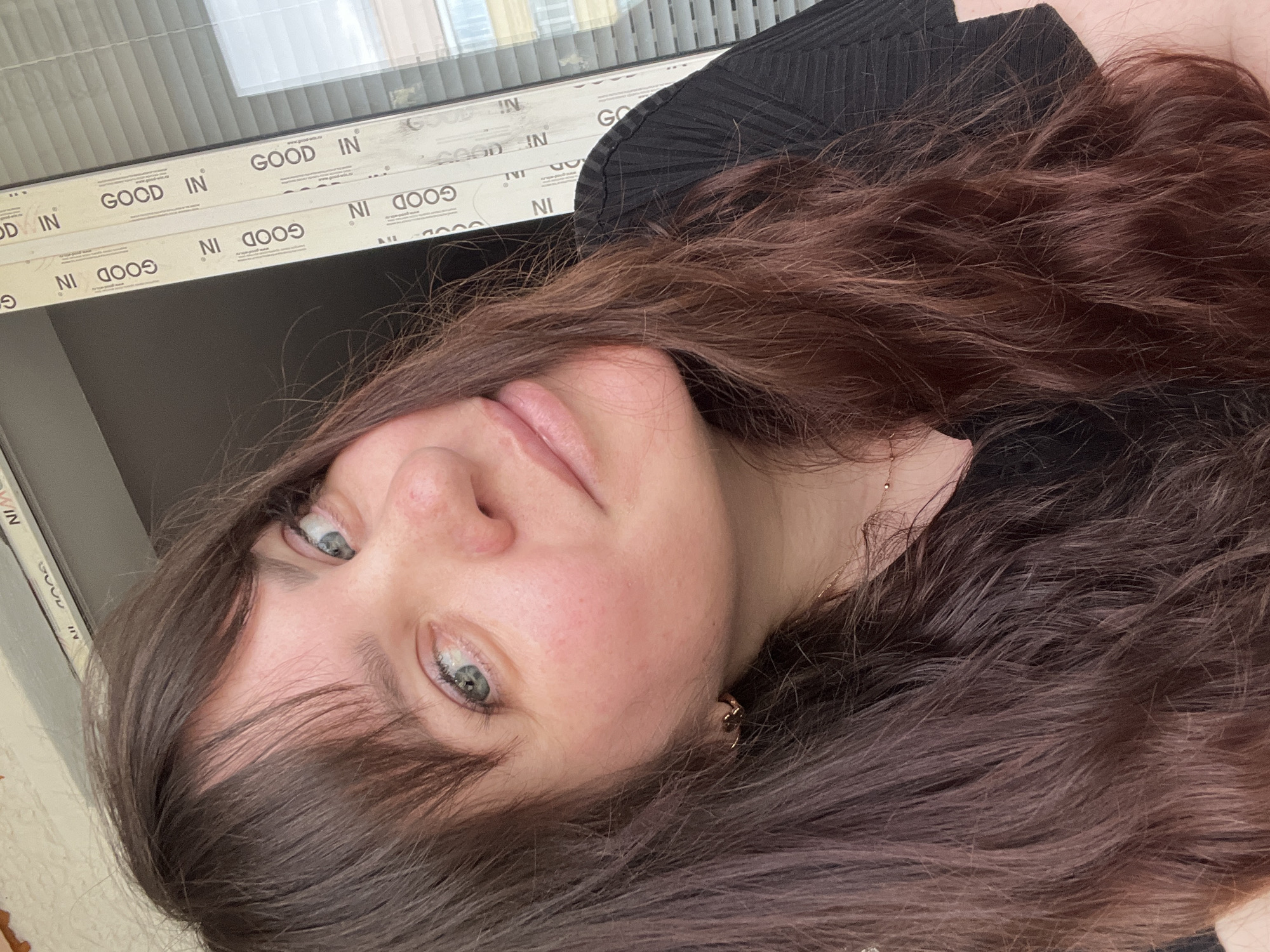 A close-up, self-taken photograph shows a young woman with brown hair and light blue eyes. Her head is tilted slightly to the right, and she is looking towards the viewer with a soft expression. Her hair is long and wavy, falling over her shoulders. She is wearing a black top and a small gold earring in her left ear. The background appears to be a window with a blind, and a strip of tape with the text "GOOD IN" printed repeatedly. The lighting suggests an indoor setting, possibly near a window. The backdrop also includes a glimpse of buildings in the distance. The photograph's perspective indicates it was taken by the subject herself.