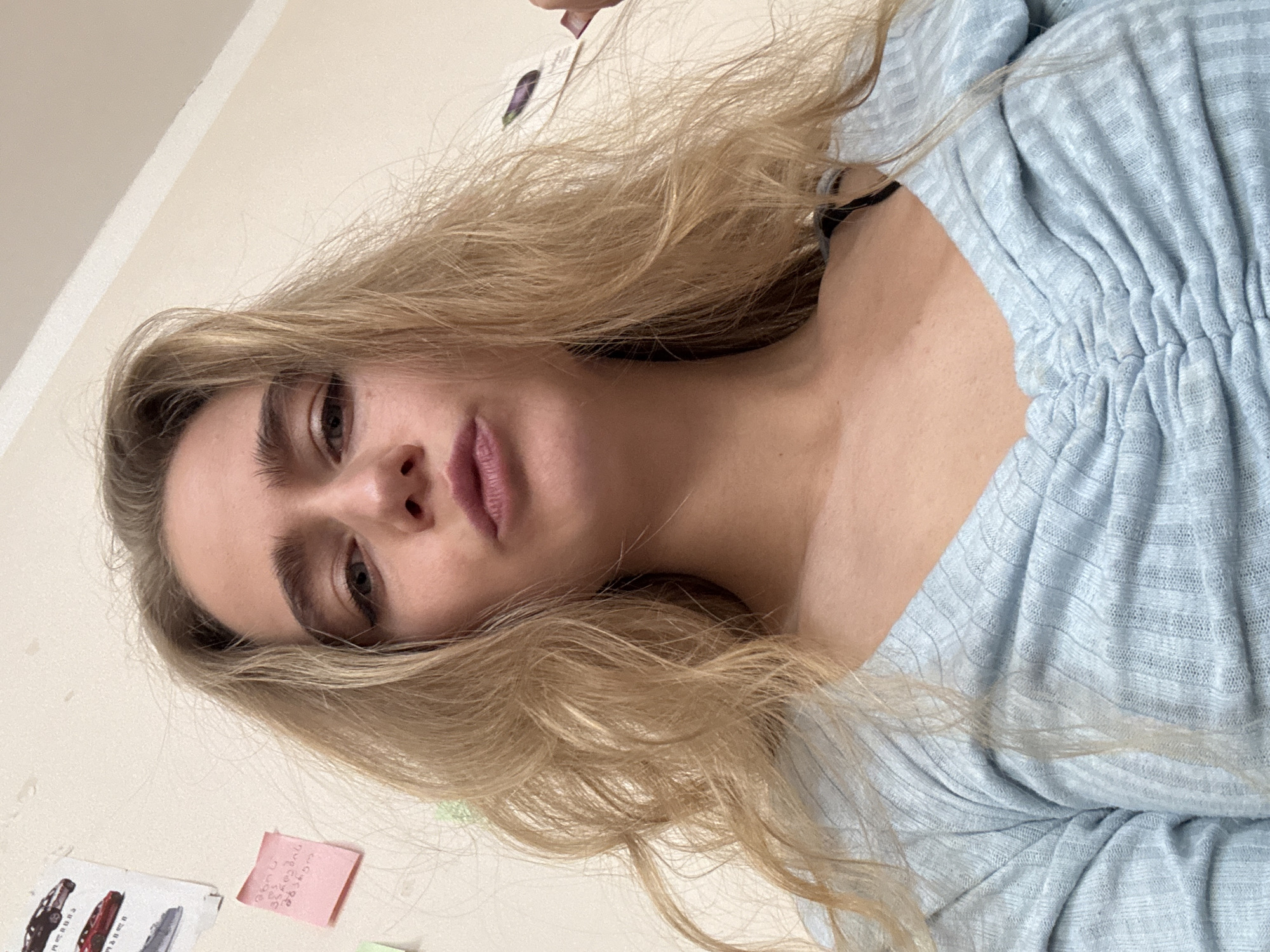 A blonde-haired individual, possibly female, is depicted in a close-up, tilted shot. The person is wearing a light blue, gathered-front garment. The background features an off-white wall and a collection of small, colorful notes or cards. The location appears to be indoors. The city is Unknown, Unknown.