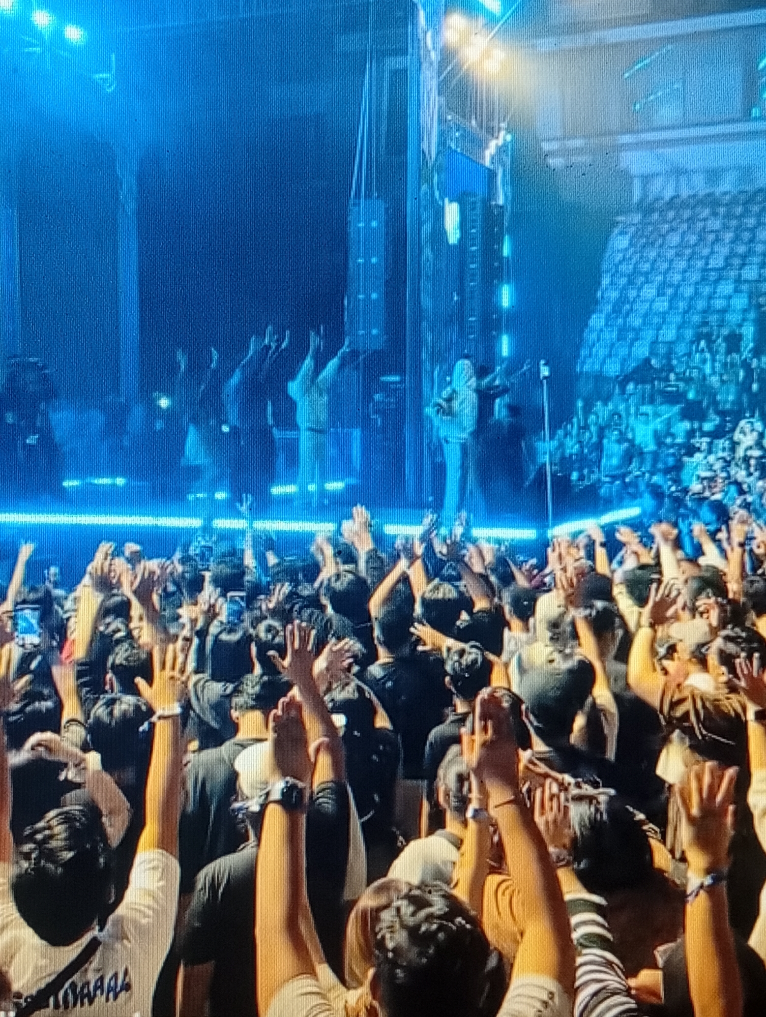 This media file captures a vibrant concert scene, likely in Lhokseumawe, Indonesia, illuminated by a wash of blue stage lighting. The primary subjects are a large crowd of people packed together, all enthusiastically engaged with the performance. Their arms are raised, many holding up cell phones to capture the moment, indicating a lively and interactive atmosphere.
The setting is clearly a concert venue, with a stage visible in the background. On stage, figures are performing, though they are somewhat obscured by the lighting and distance. Large speaker systems are prominent, suggesting a significant sound setup for the event. The crowd appears diverse in age, though predominantly young, and their raised hands and upright postures suggest excitement and participation.
The activity is a live musical performance, with the audience actively responding to the music and the performers. The time of day is not explicitly indicated, but the intense artificial lighting implies an evening or indoor event. There are no discernible weather cues.
Notable details include the overwhelming presence of cell phones being used to record the concert, a common sight at modern events. The overall emotion conveyed is one of exhilaration and shared enjoyment. There is some visible text on a person's shirt in the lower left quadrant, appearing to be stylized lettering, possibly a logo or band name, but it is not clearly legible.