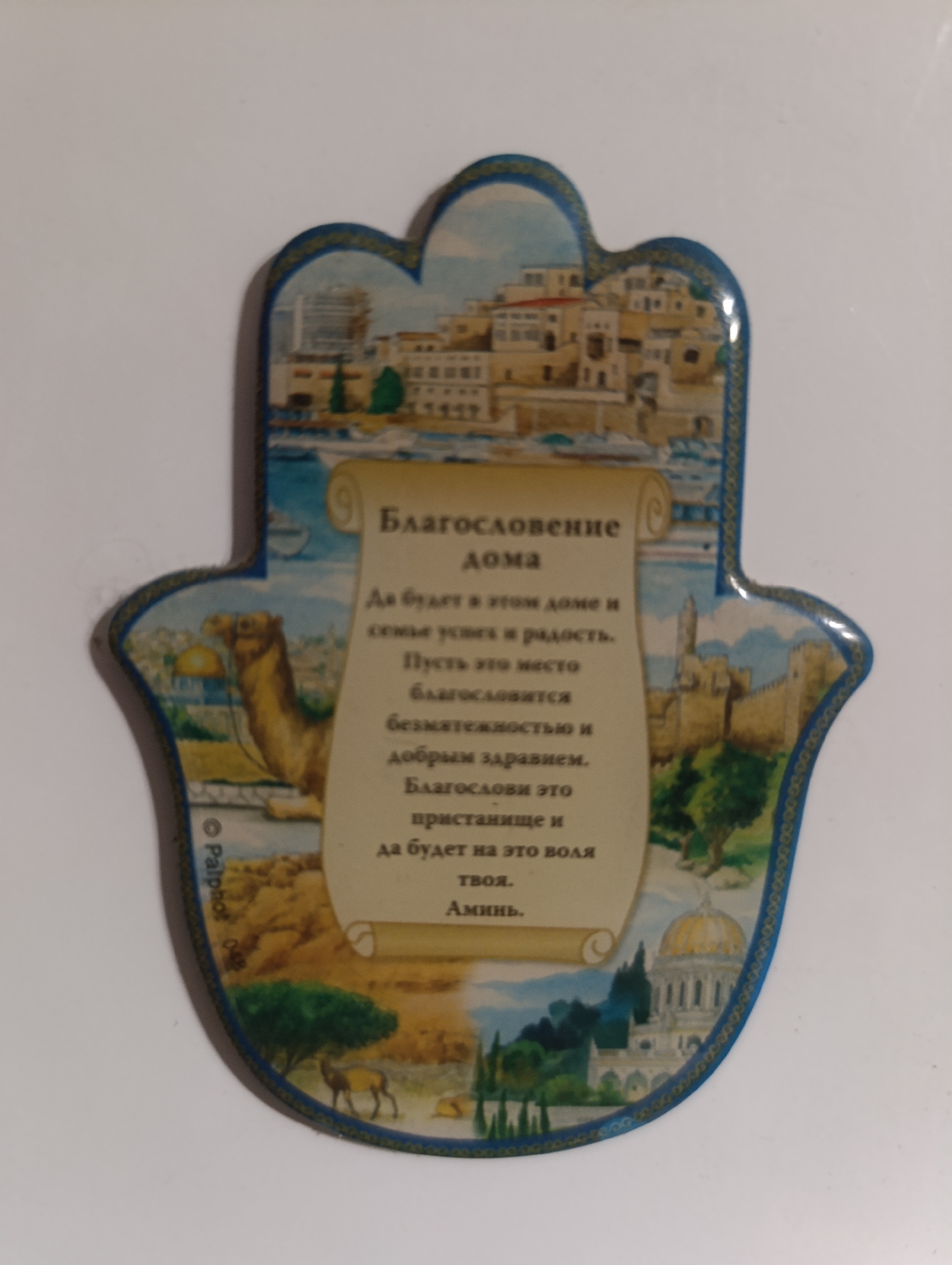 A hamsa, or hand of Fatima, amulet featuring a painted scene of an ancient city with buildings and a camel, and a scroll with text. The text appears to be in Russian and translates to "Blessing for the Home: May there be in this home, family unity and joy. May this place be blessed with abundance and good health. May blessing and solace be upon this home. Thy will be done. Amen." The amulet is placed on a white surface.