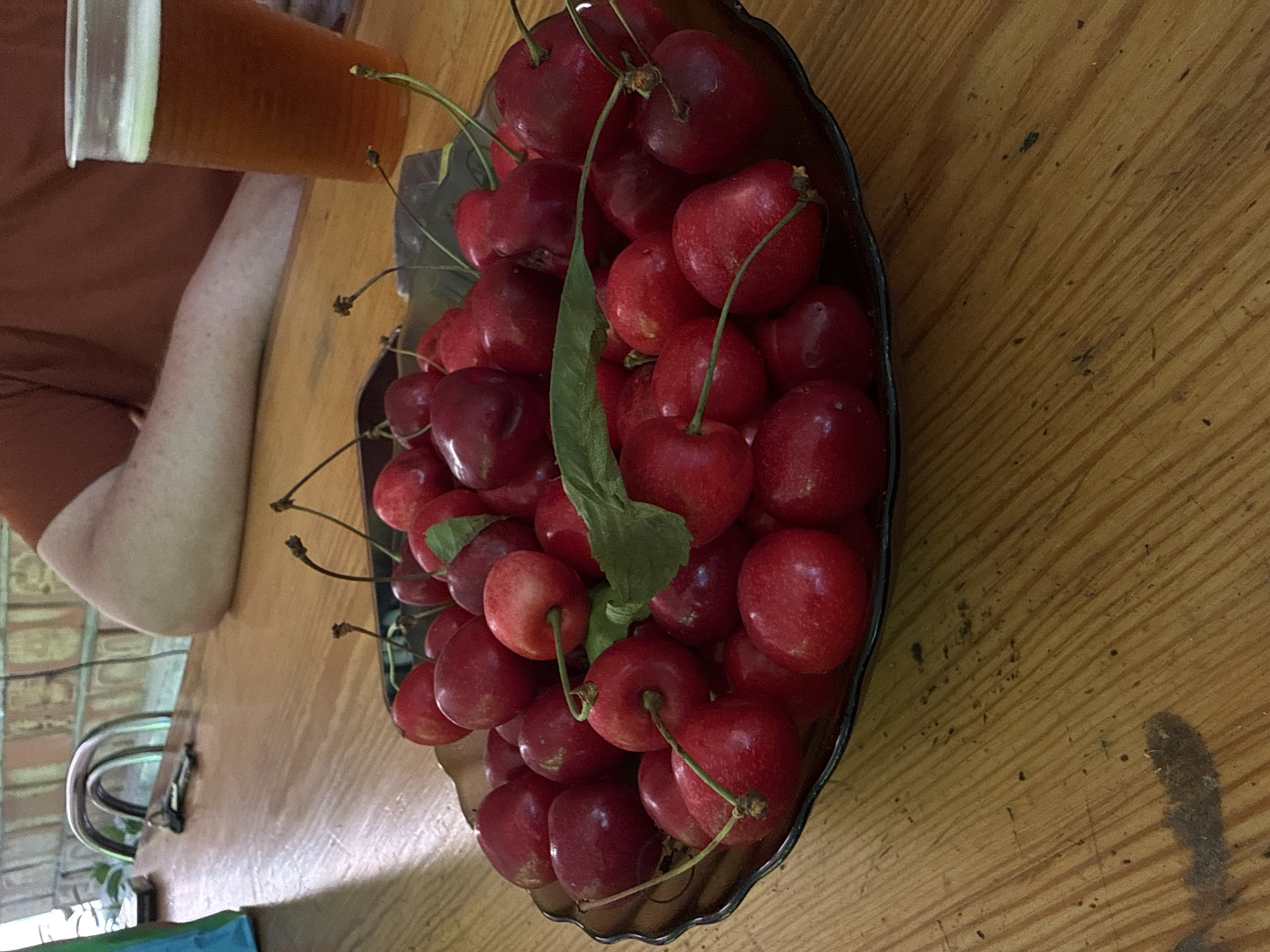 A close-up, eye-level shot showcases a dark, scalloped-edge bowl filled with fresh, plump red cherries. The cherries are piled high, some with stems still attached, and a few green leaves are scattered among them. The bowl rests on a light-colored wooden table with visible grain and some dark smudges. In the background, a person's arm and shoulder are visible, wearing a reddish-brown shirt. To the right of the cherries, a clear plastic cup contains a amber-colored liquid, likely beer, with a frothy white head. The environment suggests a casual outdoor or indoor setting, possibly a cafe or a home patio, in Obukhivka, Ukraine.