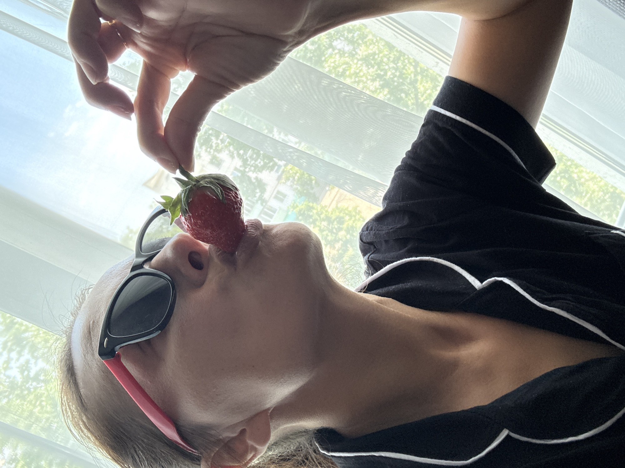 A woman with sunglasses is holding a strawberry to her mouth. She is wearing a black shirt with white trim. The location is possibly in the city of Chaikovsky, Russia, given the request. The background shows a window with a blurred view of trees.