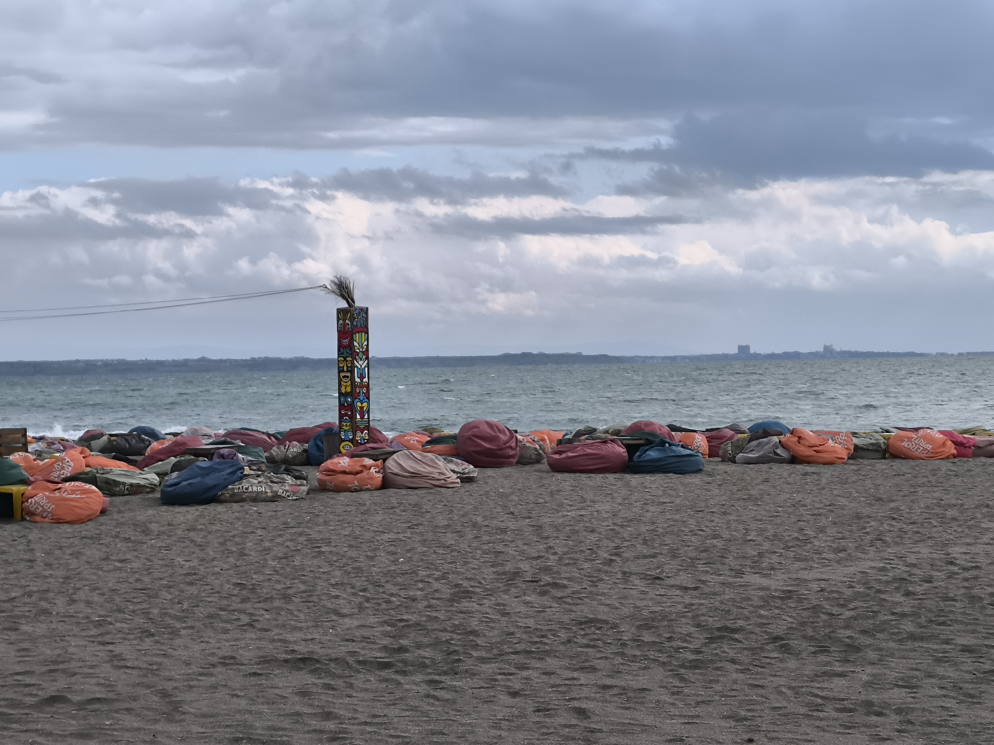 The image depicts a beach scene in Burgas, Bulgaria. A dark sand beach fills the foreground. A row of colorful beanbag chairs, branded with names like "Aperol Spritz" and "Bacardi," runs across the midground. Behind the chairs is the Black Sea, with small waves. On the left, there is a wooden structure partially visible, while a totem-like pole stands out near the center. The pole is adorned with painted faces and topped with a small tuft of reeds or straw. The sky is overcast with grey and white clouds. A distant landmass is visible across the water, suggesting the coastline or a distant part of Burgas. No people are immediately apparent in the shot.