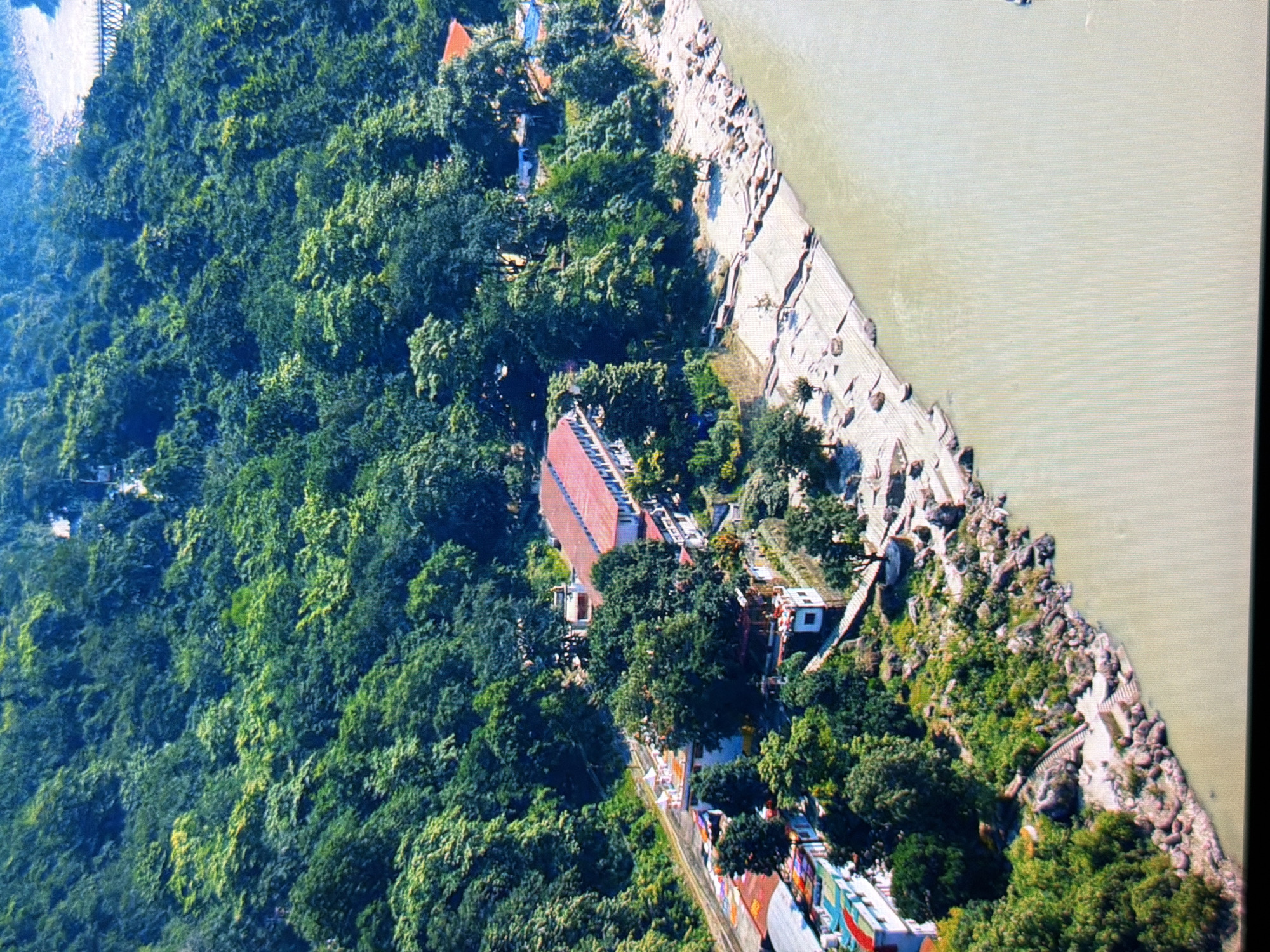 An aerial view captures a lush, green forested hillside bordering a wide, muddy river. Several buildings with red and white roofs are nestled amongst the trees on the hillside, some appearing to be part of a resort or spiritual center, with a road running alongside them. The riverbank is rocky and has some sandy patches, with steps leading down to the water. A suspension bridge is visible in the distance on the upper right side of the image. The overall scene depicts a tranquil natural setting with man-made structures integrated into the landscape. This image is not located in Kyiv, Ukraine.