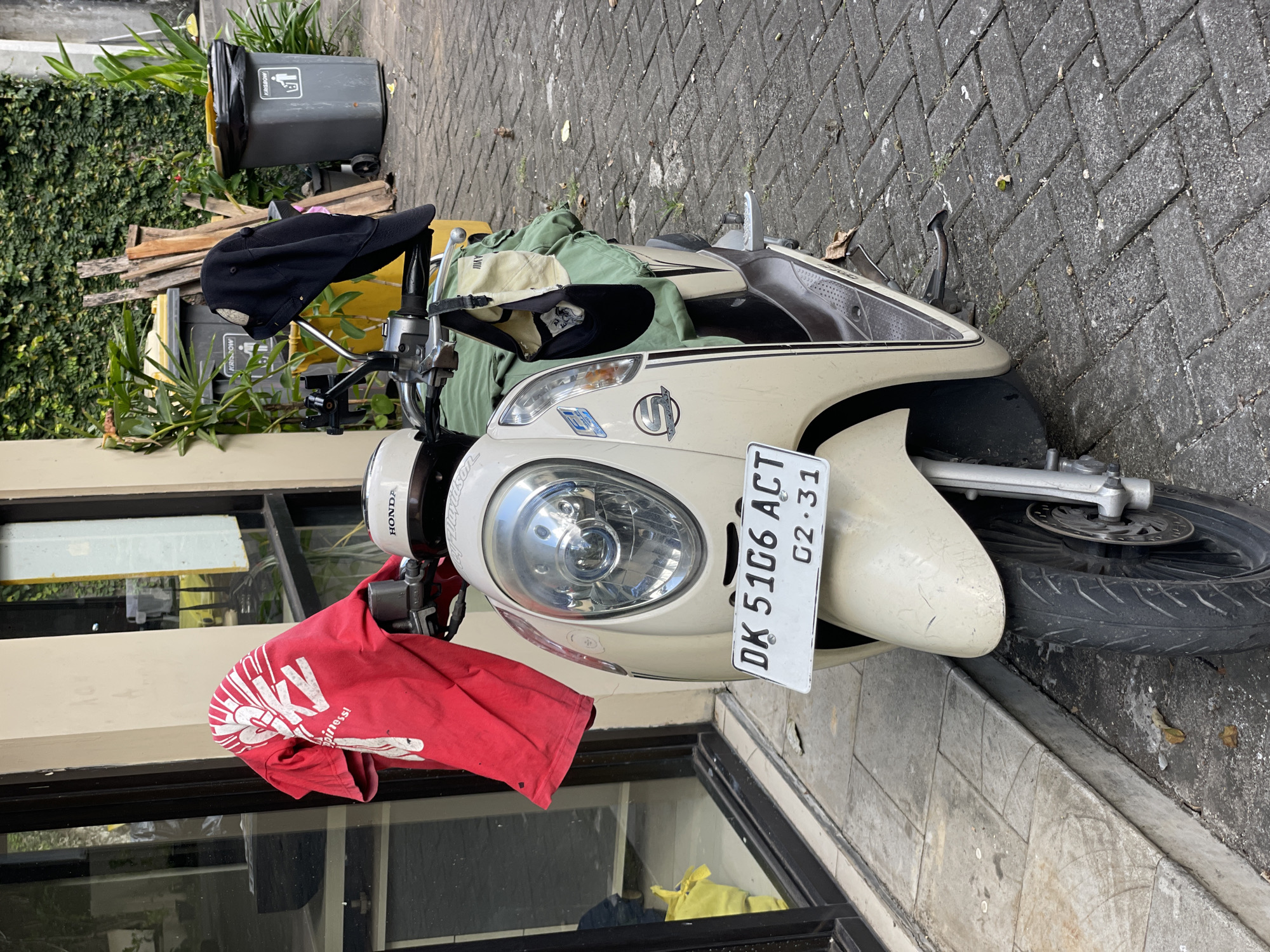 A beige Honda scooter with the license plate "DK 5106 ACT" is parked on a brick-paved sidewalk. A red t-shirt with a white print is draped over the scooter's handlebars, while a black baseball cap and a tan baseball cap are resting on the scooter's body. Behind the scooter, a gray garbage bin is visible, along with some pieces of wood. A building with glass windows and a green wall of foliage is also in the background. The motorcycle license plate indicates that the location is in Kuta Selatan, Indonesia.