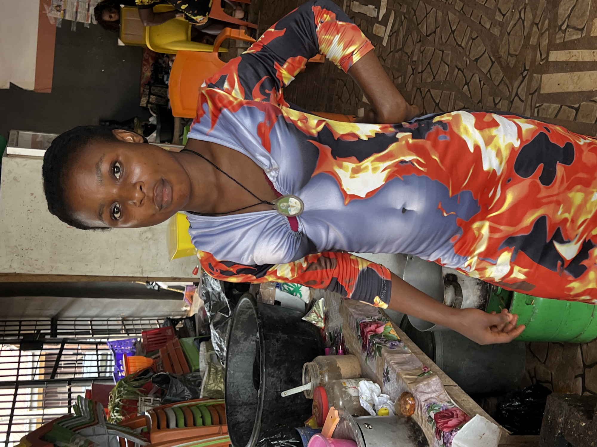 A young Black woman stands in what appears to be a marketplace or shop in Nkpor, Nigeria. She is wearing a long-sleeved, form-fitting dress with a vibrant flame-like pattern in shades of red, orange, yellow, and black on the sleeves and skirt, transitioning to a light blue on the bodice. A dark cord necklace with a circular pendant containing a picture is visible around her neck. Her left hand rests on her hip.
The background is cluttered with various items. To the left, stacked colorful plastic chairs (orange, green, and white) are piled high, along with other market goods like plastic containers and bags of what could be produce or supplies. A large black basin or tub is prominent on a table draped with a floral-patterned cloth. Jars containing powders and other items are also on the table. Further back, the environment includes concrete walls, metal bars resembling a security grate, and other individuals in the distance, including a child sitting on a yellow chair. The floor is tiled with a stone pattern. The lighting suggests an indoor or covered outdoor space.