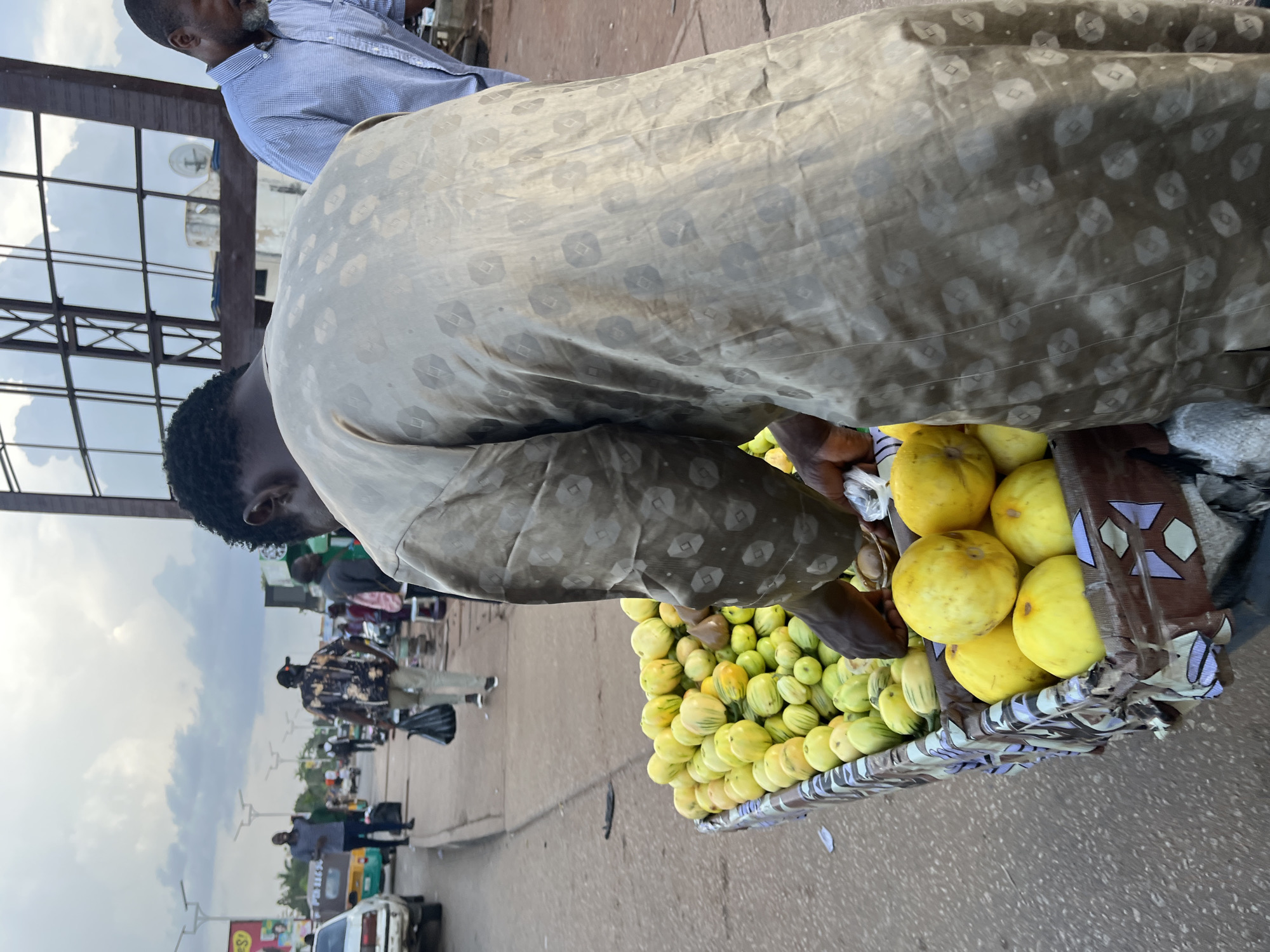 A man in a patterned tan tunic stands behind a cart overflowing with yellow-green and yellow produce, likely fruits or vegetables, on a street in Barakin Lerawi, Nigeria. The cart is adorned with a patterned fabric. To the right of the fruit vendor, another man in a blue checkered shirt is visible. In the background, several people walk along a paved sidewalk next to a road where vehicles, including a white car and a green and yellow auto-rickshaw, are present. Buildings with glass facades and utility poles line the street. The sky is overcast.