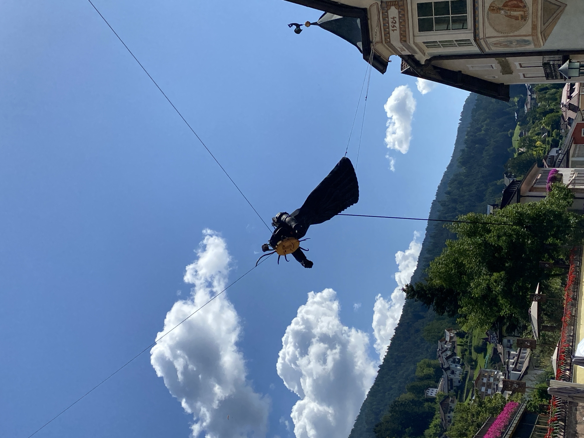 A puppet with a sun-like face and black, flowing garment is suspended by wires against a bright blue sky with scattered white clouds. The puppet is positioned in the foreground, slightly to the left of the center. In the background, a steep, tree-covered mountain rises, dotted with houses. The town of Urtijëi - St. Ulrich in Gröden - Ortisei, Italy is visible on the mountainside, with traditional alpine architecture. A building with a decorative facade and the year "1984" is visible to the right of the puppet.