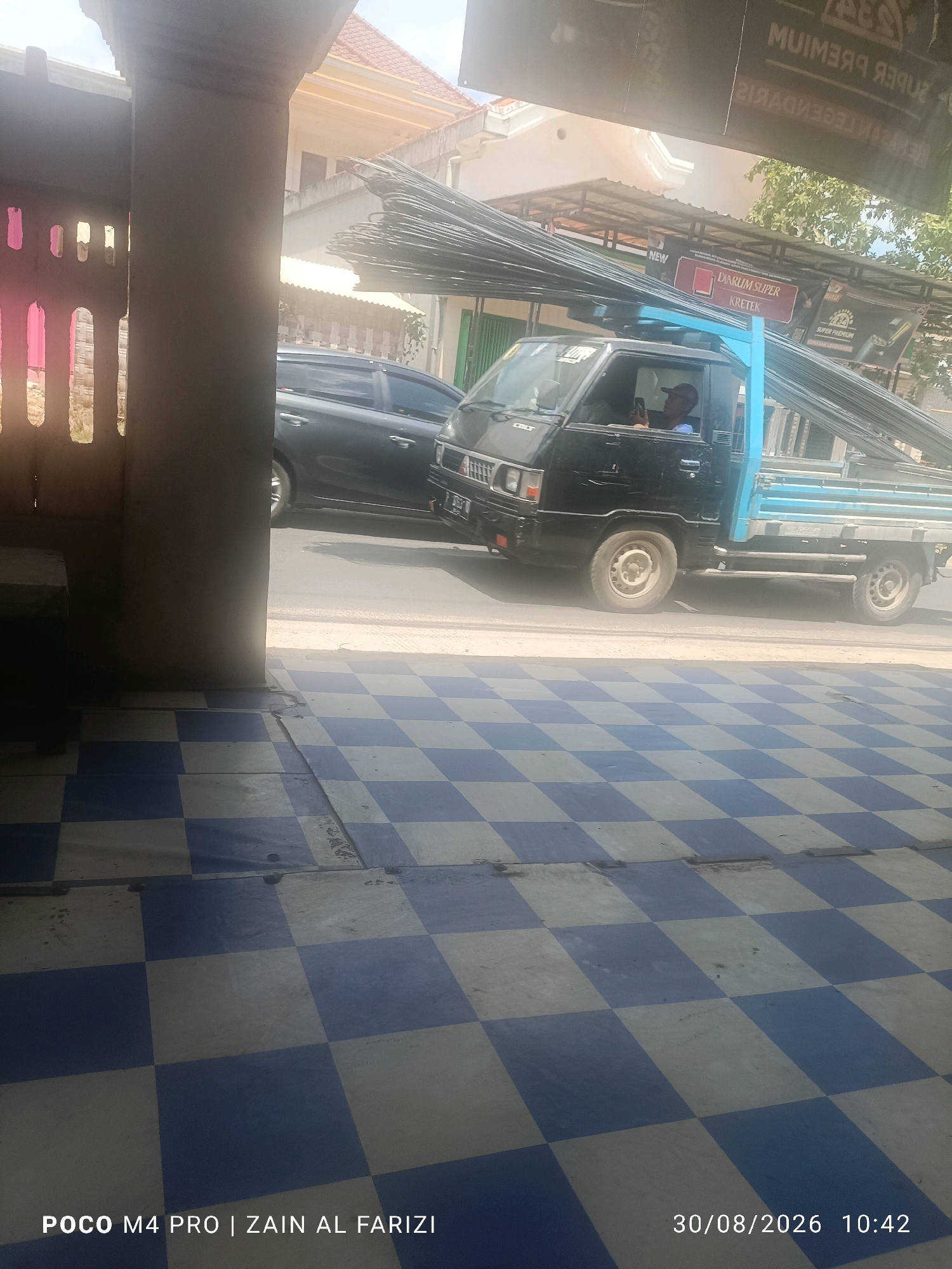 In the city of Karang Pao, Indonesia, a black Mitsubishi L300 pickup truck carrying a large load of metal rebar is parked on a street. The truck is positioned to the right of a concrete pillar and the foreground features a checkered tile floor in blue and off-white. In the background, a silver sedan is visible on the street. A man wearing a dark cap is seated in the driver's seat of the truck, holding a mobile phone. Above the truck, a banner with text, some of which is legible, hangs across the street. The scene is illuminated by natural daylight.