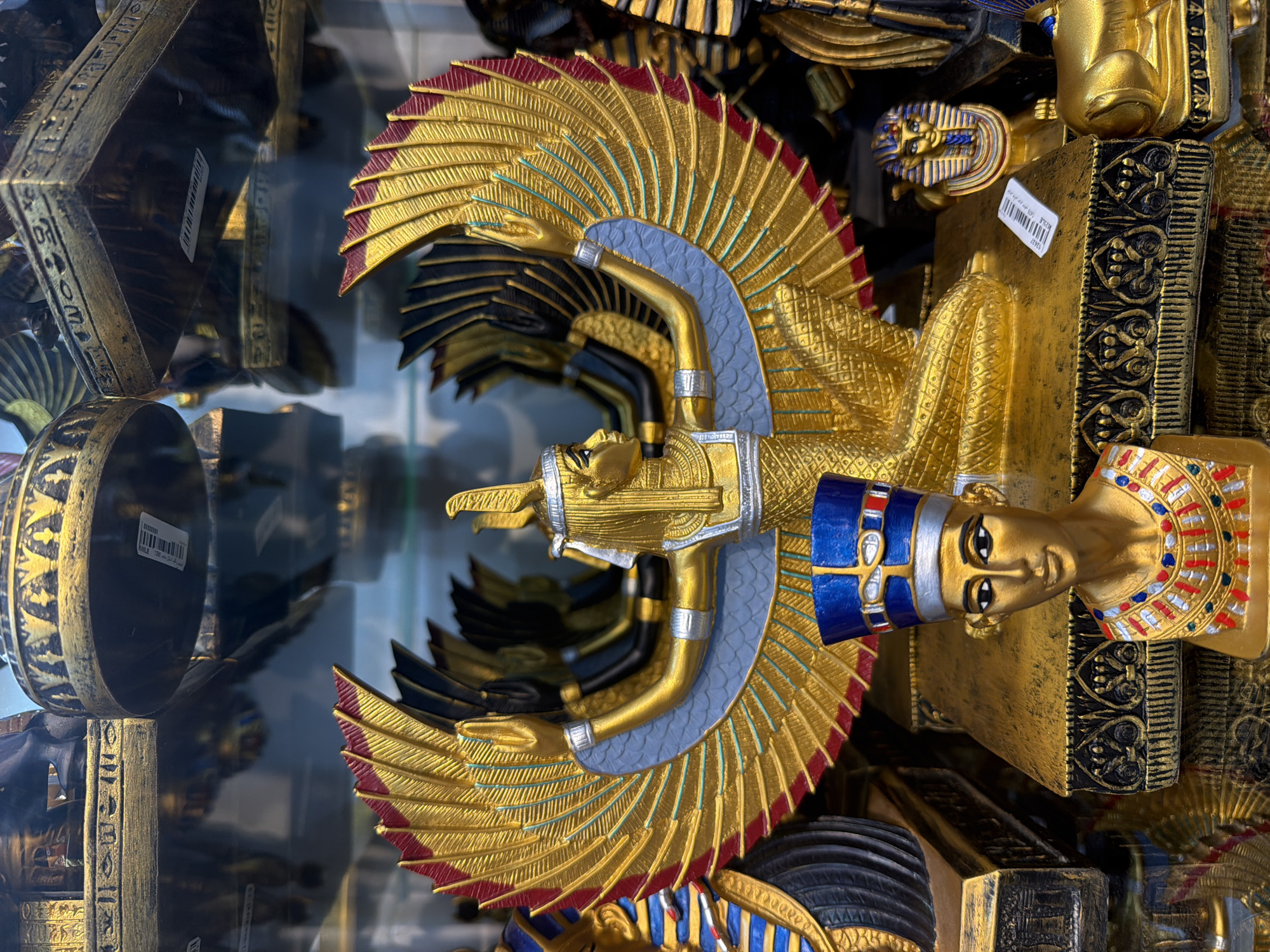 The image shows a close-up view of Egyptian-themed souvenirs displayed on shelves, likely in a shop. The primary subject is a large golden statue of a winged goddess, possibly Isis or Nephthys, with her arms outstretched and wings spread. In front of her is a smaller bust of Nefertiti wearing her iconic blue headdress. Several other Egyptian artifacts, including miniature sphinxes and decorative elements with hieroglyphics, are visible in the background, some of which appear to be reflected in glass surfaces. The lighting suggests an indoor setting, possibly a retail environment in Sharm El Sheikh, Egypt, given the context. The overall impression is of a collection of decorative items related to ancient Egyptian culture.