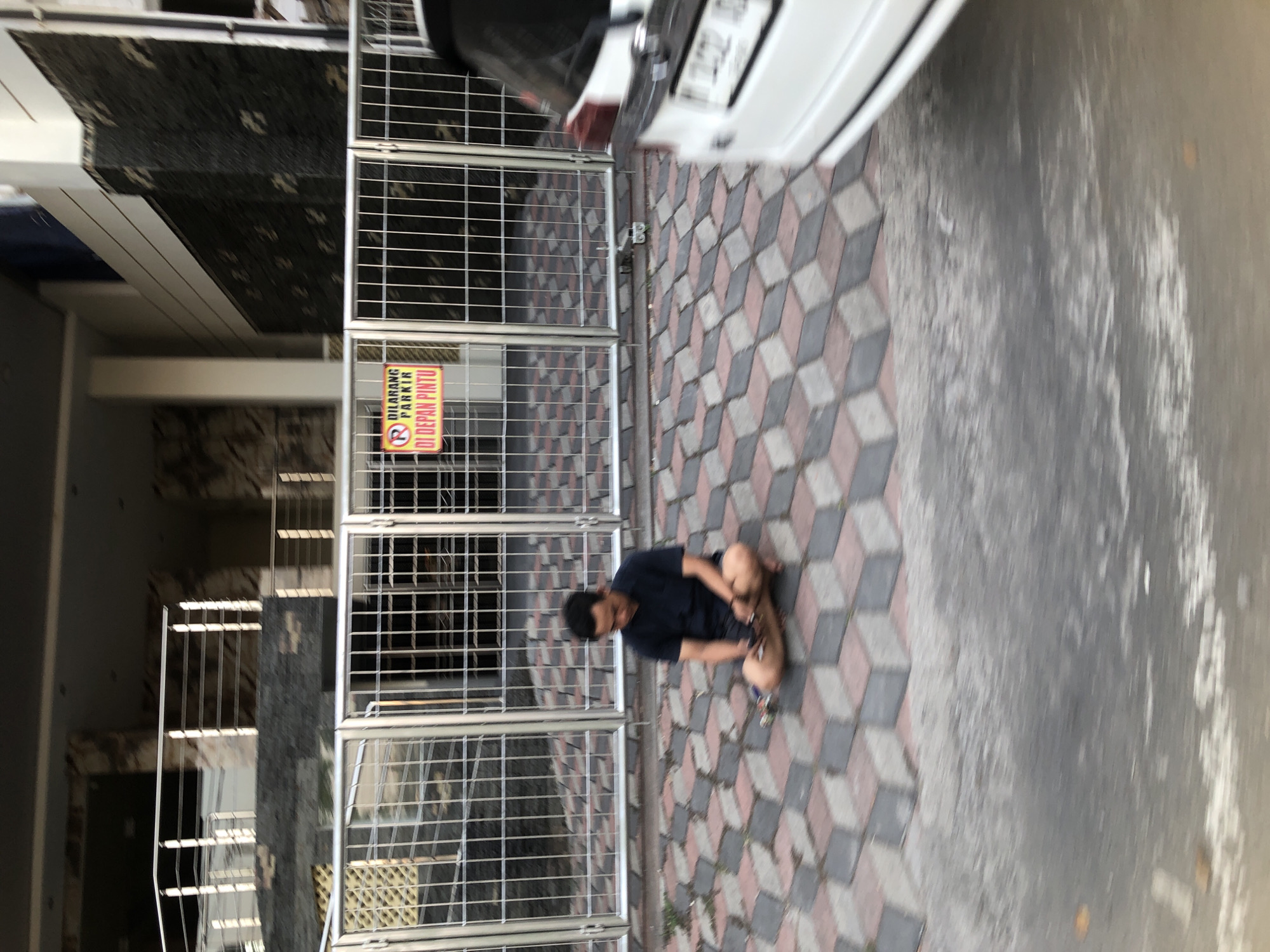 A full-shot vertical image shows a man sitting cross-legged on a tiled patio. The man is wearing a dark blue short-sleeved collared shirt and tan shorts. He is looking down at his hands, which are resting on his lap. To the left of the man is a metal gate with a yellow and red sign that reads "DILARANG PARKIR DI DEPAN PINTU" (No Parking in Front of the Door). Behind the gate is a building with a stone facade. To the right of the image is the rear of a white SUV. The patio tiles are arranged in a repeating pattern of grey and reddish-brown cubes that create an optical illusion of depth. The overall scene suggests a public area, possibly near a residential or commercial building. The image was taken in Candi, Indonesia.