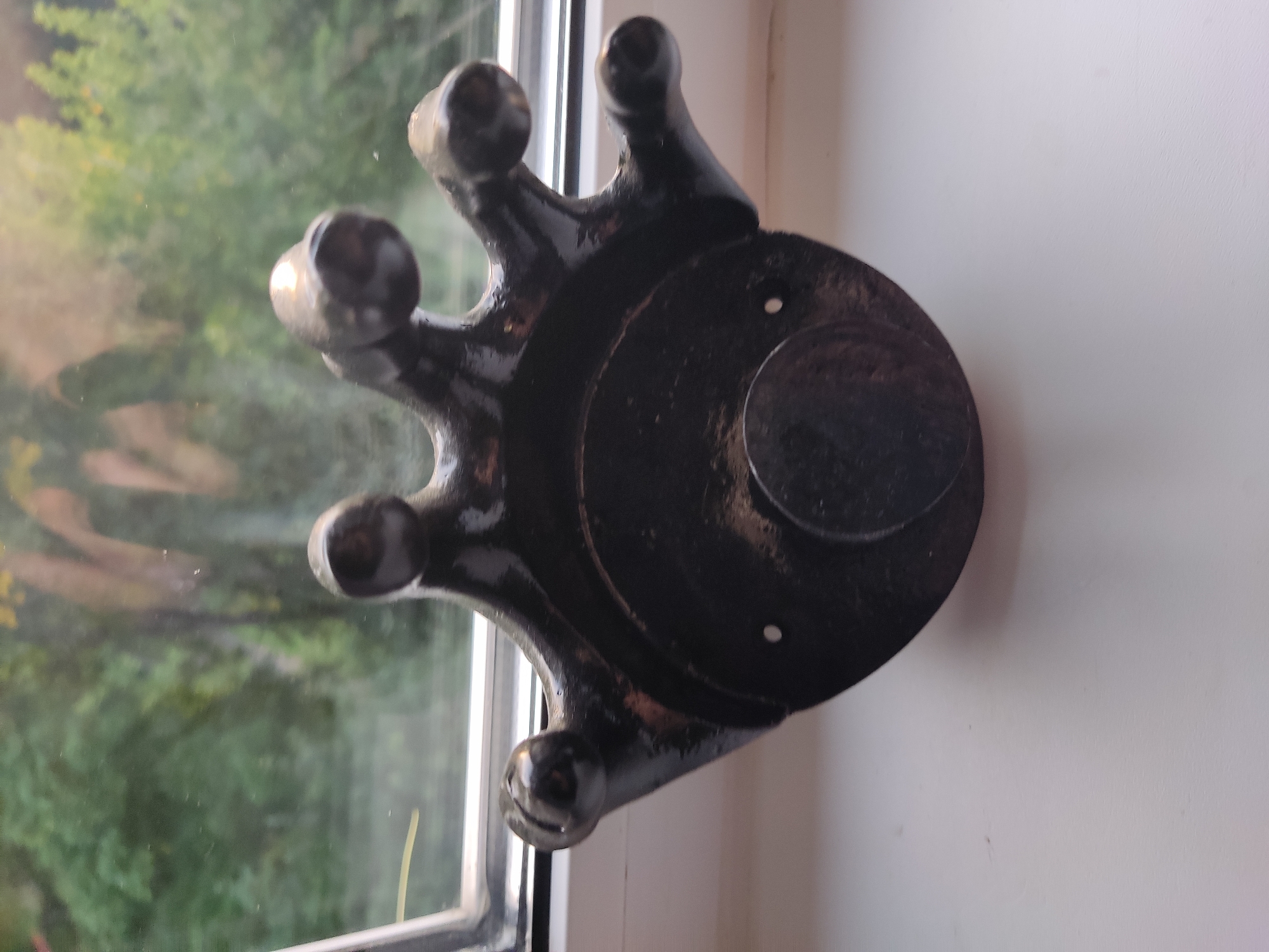 A dark, possibly metal, object resembling a multi-pronged creature or a stylized hand, sits on a white windowsill. The object has a central circular base with a smaller circular protrusion in the center. Five "fingers" or prongs extend upwards and outwards from the top of the base, each ending in a rounded knob. The object is glossy and reflects light. Behind it, a window shows a blurred view of green trees and foliage. The object is positioned in the foreground on a white surface that could be a windowsill. The image does not contain any information that would indicate a specific location in Noginsk, Russia.