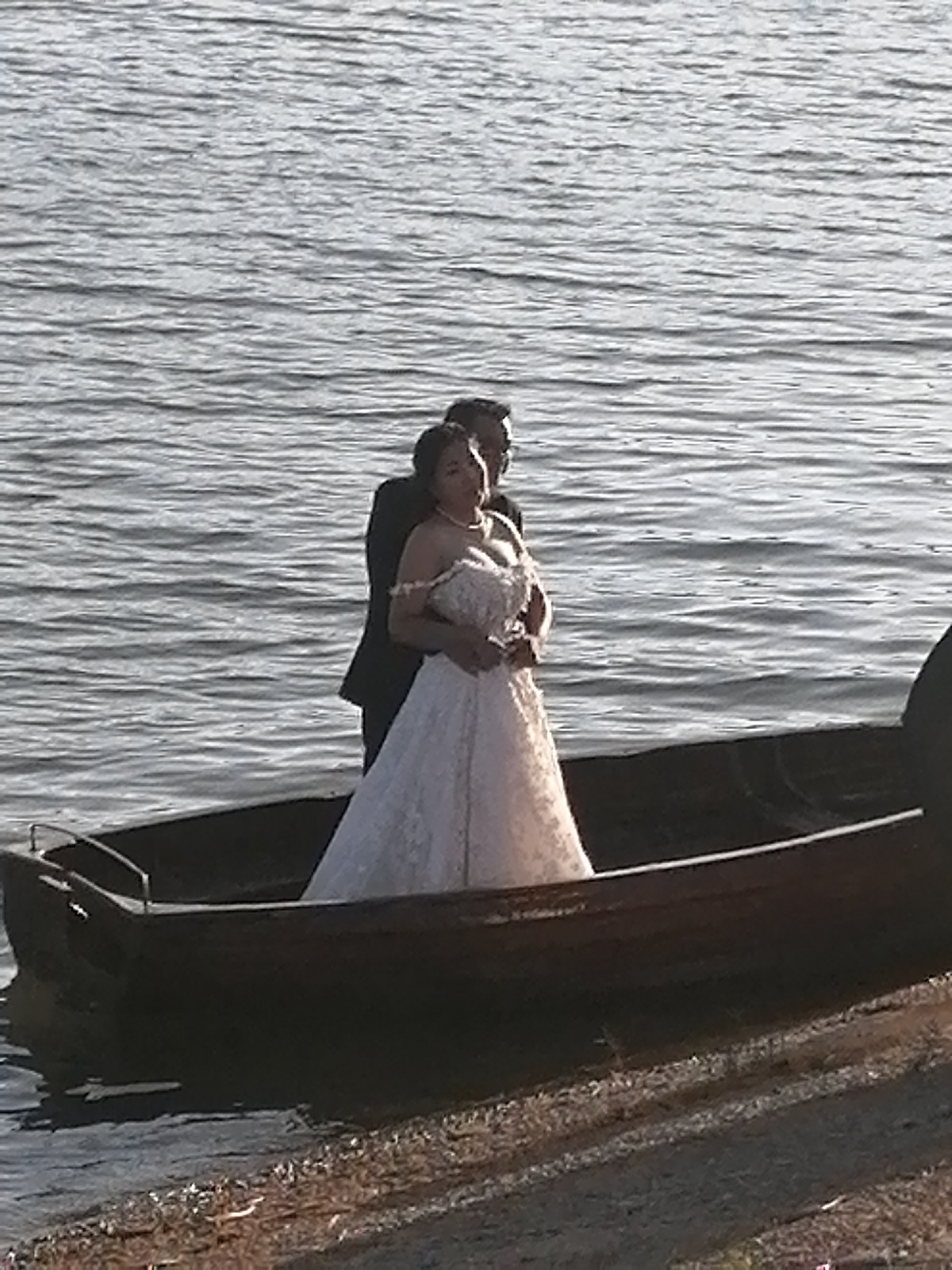 A couple stands in a boat on the water. The man is wearing a suit and tie, and the woman is wearing a white wedding dress. They are embracing, and the woman is holding a bouquet of flowers. The boat is dark in color and appears to be made of wood. The water is calm and reflects the sky. The background is out of focus but appears to be a natural setting. The image is taken from a slightly elevated angle.