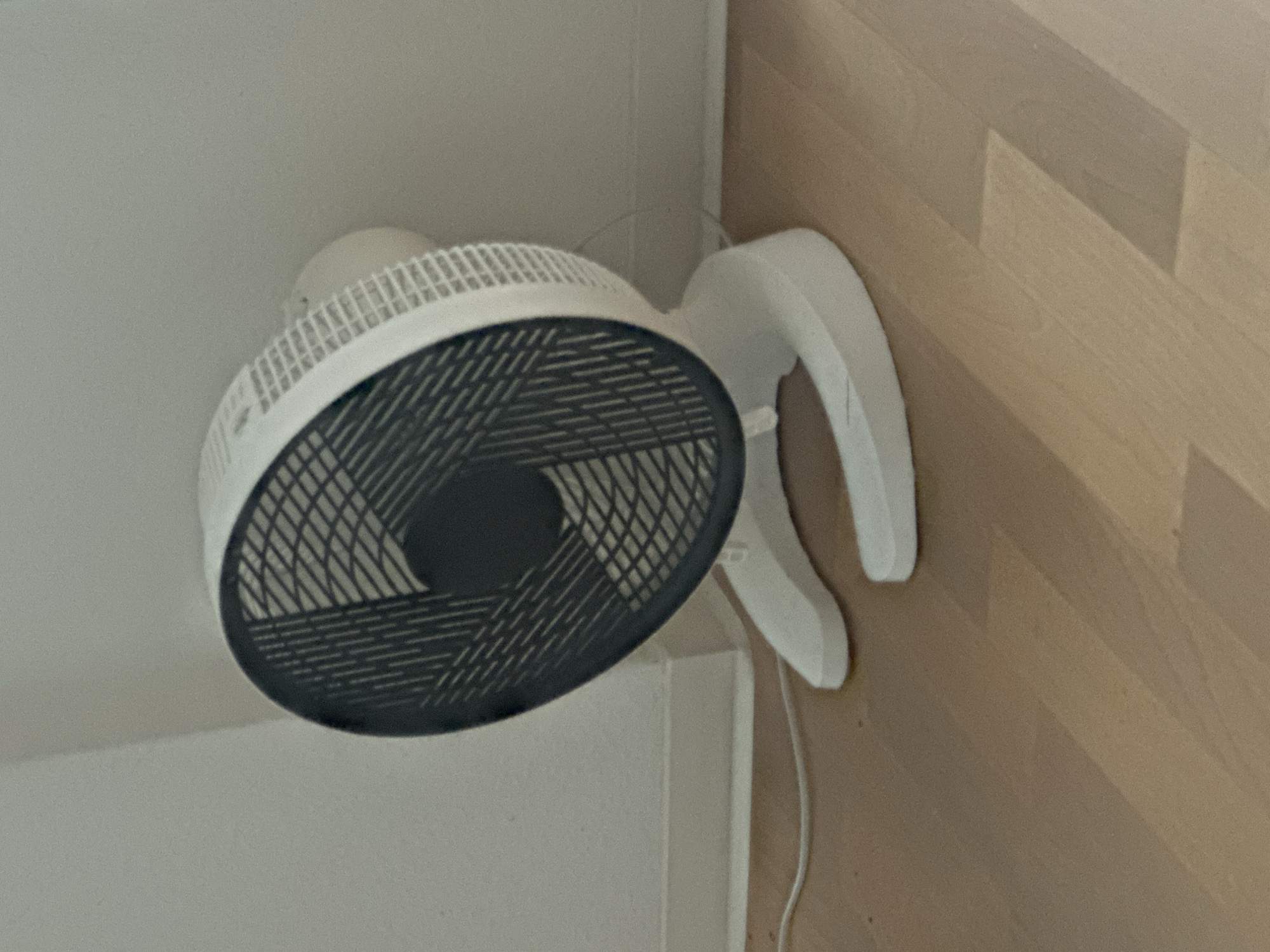 The image shows a white electric fan with a black grill, placed on a light-colored wooden floor. The fan is positioned against a white wall, with the base resting on the floor. A white power cord is visible trailing from the fan. The lighting suggests an indoor environment, possibly a room. There are no people or any other distinguishing features visible in the image. The scene is entirely indoors, and there is no visible indication of the location being in the city of Görlitz, Germany.