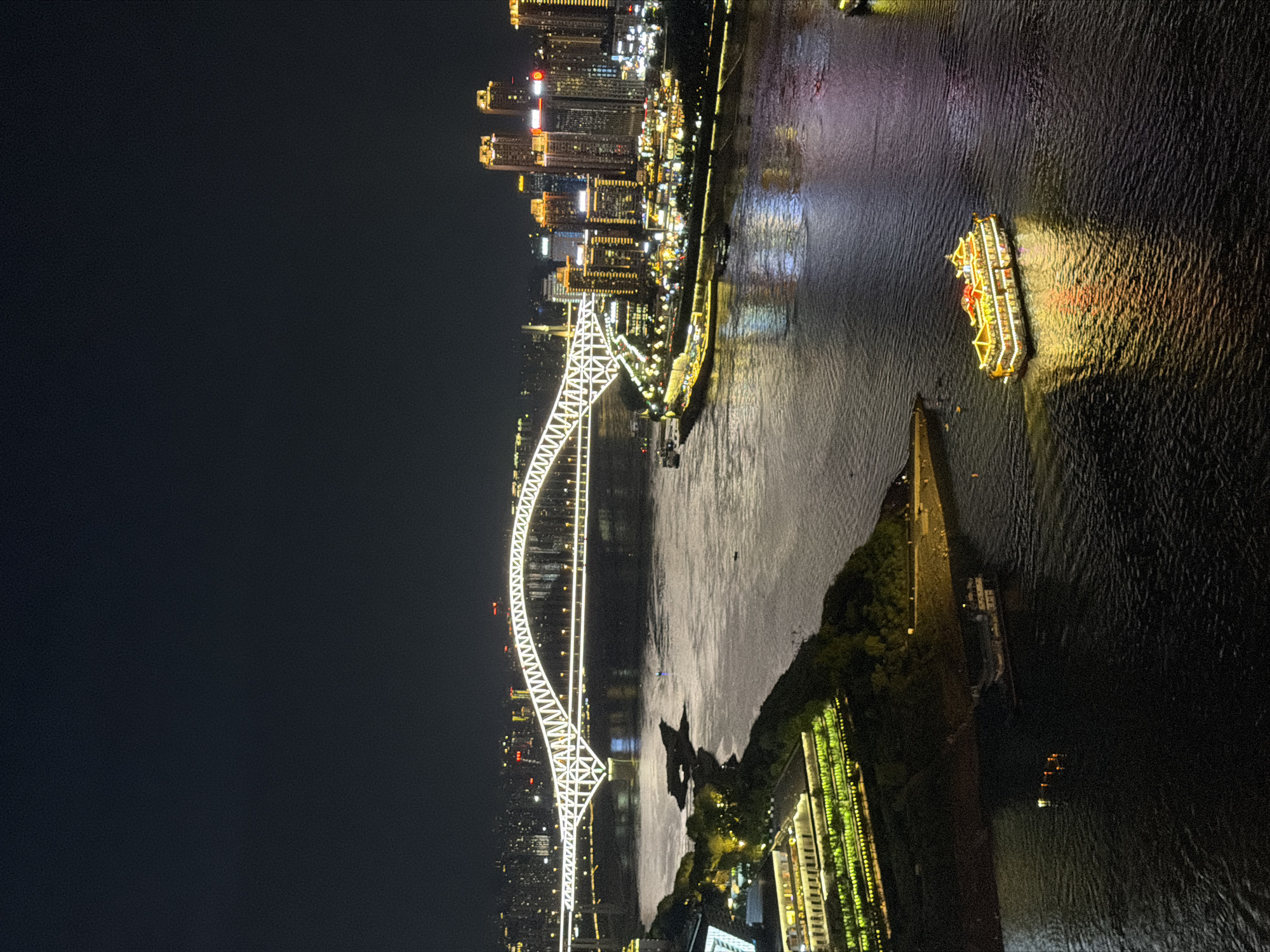 An aerial, nighttime view captures the Yuzhong District in Chongqing, China, dominated by the brightly illuminated Yangtze River Bridge spanning a wide, dark river. The bridge, a prominent white structure with intricate geometric patterns, is the central focus, its lights reflecting on the water's surface.
On the left bank, a multi-story building with extensive green terracing and numerous yellow lights illuminates the foreground. Adjacent to this, a darker, sloped embankment leads down to the water, with trees and some scattered lights visible. Further into the river, a large, ornate boat with a yellow roof and multiple levels of illuminated windows is a significant subject, its bright lights casting a strong reflection on the dark water. Several smaller boats are also present, some with lights and others appearing as dark shapes.
The right bank of the river is a densely built-up urban landscape, characterized by numerous tall skyscrapers of varying heights. These buildings are illuminated with a mix of yellow and white lights, creating a vibrant city skyline that stretches into the distance. The overall impression is one of a bustling, modern city at night, with the illuminated bridge and the river serving as a dramatic backdrop. The sky is a deep, dark black, with no stars or moon visible.