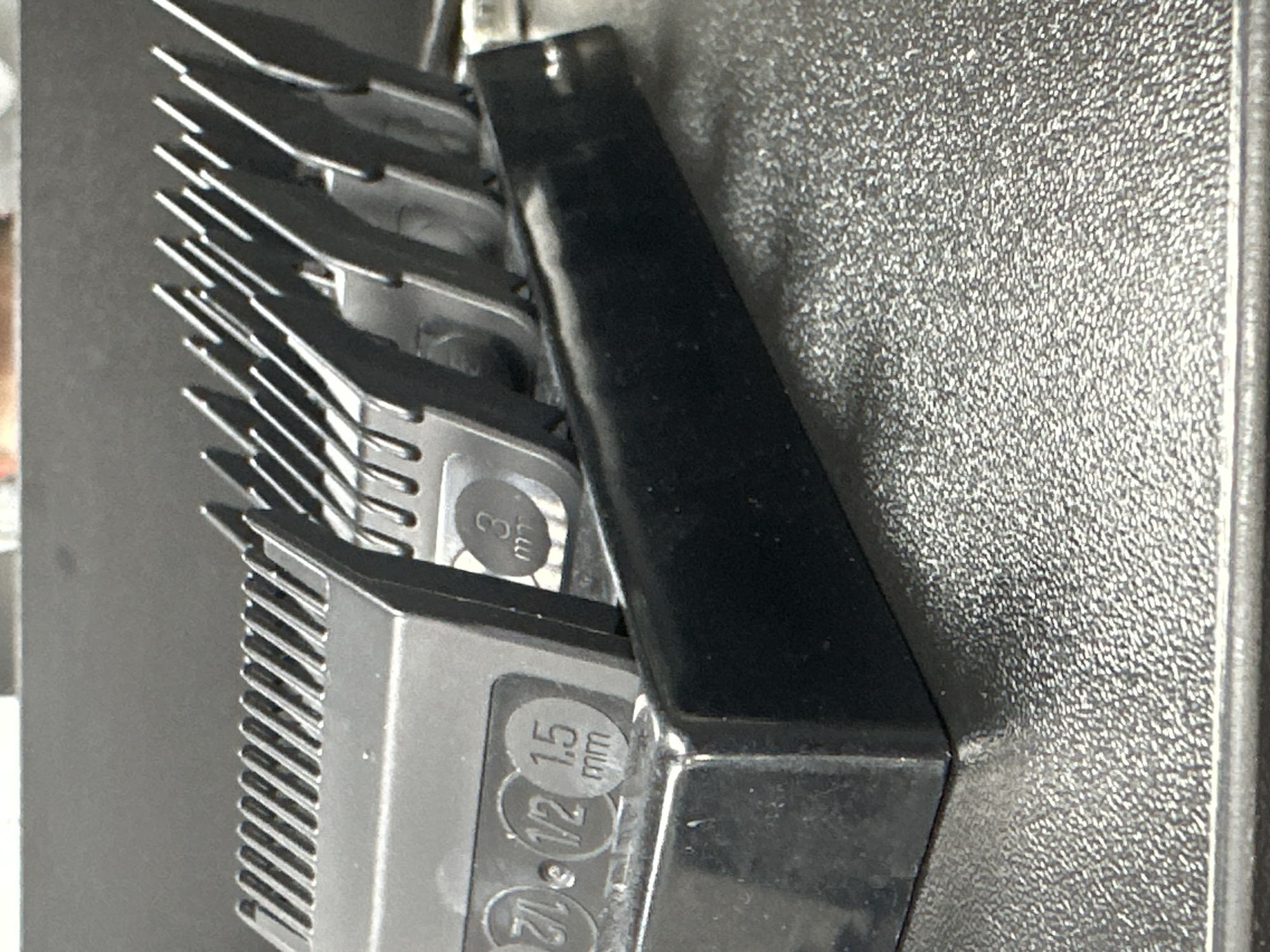 A black plastic stand holds several metal hair clipper guards. The guards are labeled with measurements: 2/1, 1/2, 1.5 mm, and 3 mm. The stand is placed on a textured grey surface. The background is blurred and dark. This image does not depict any location in Uzhhorod, Ukraine.