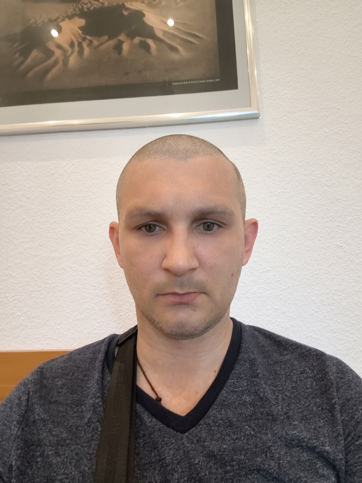A man with a shaved head and a dark blue V-neck shirt is photographed from the chest up. He has light skin, green eyes, and a faint shadow of stubble on his chin and jawline. A dark strap is visible across his chest, and a thin brown cord with two dark beads hangs around his neck. Behind him is a white, textured wall, and above it, a framed photograph of sand dunes. A light brown wooden surface is visible along the bottom edge of the frame. The image is taken indoors, with neutral lighting.