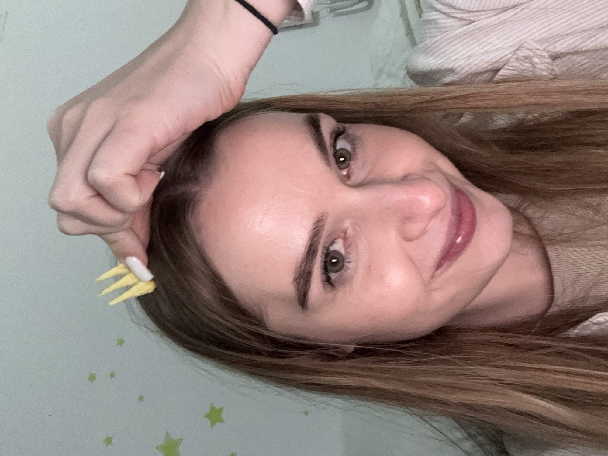 A close-up image depicts a Caucasian female, appearing to be in her mid-20s to early 30s, positioned indoors in what is described as the city of Budva, Montenegro. She has long, light brown hair, green eyes, and is smiling faintly directly at the camera. Her right hand, adorned with white-tipped fingernails, is raised above her head, holding a small, yellow, three-pronged object that resembles a miniature fork or crown. She wears a light-colored, possibly beige, ribbed top beneath a light-colored pinstriped collared garment. A black hair tie is visible on her left wrist. The background consists of a plain, light greenish-blue wall decorated with several light green star-shaped decals of varying sizes.