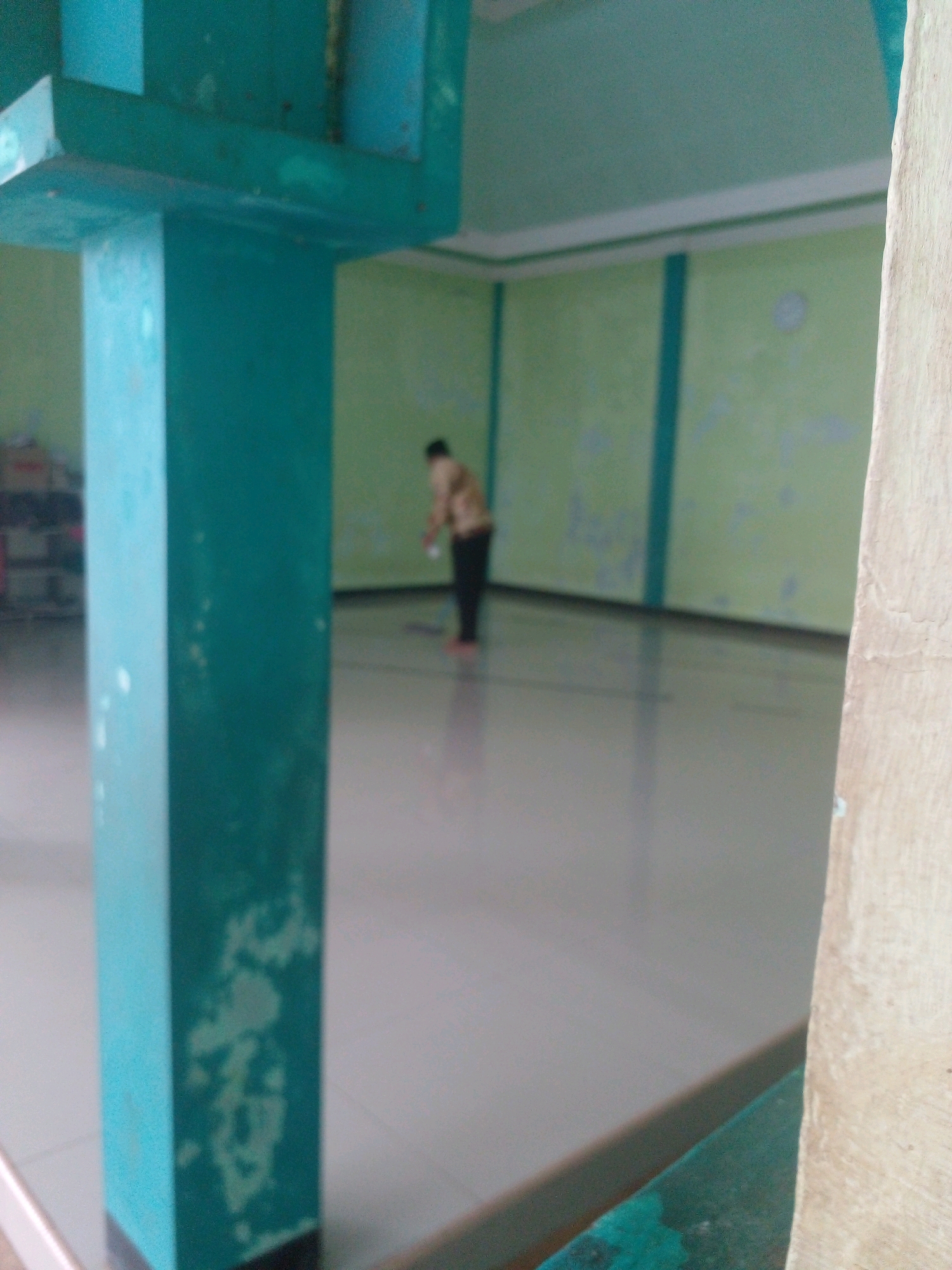 The image depicts the interior of a building, possibly a mosque or community hall, with a person sweeping the floor. A large, turquoise-colored pillar is prominently featured in the foreground on the left, showing signs of wear and discoloration. The walls of the building are painted a pale green with turquoise accents along the top and vertical stripes. The floor is tiled and appears to be reflective, showing the ambient light. In the background, a lone individual, seen from the back, is engaged in sweeping, their attire consisting of a patterned shirt and dark trousers. The overall impression is one of a functional, somewhat aged interior space. The location is Banyuputih, Indonesia.