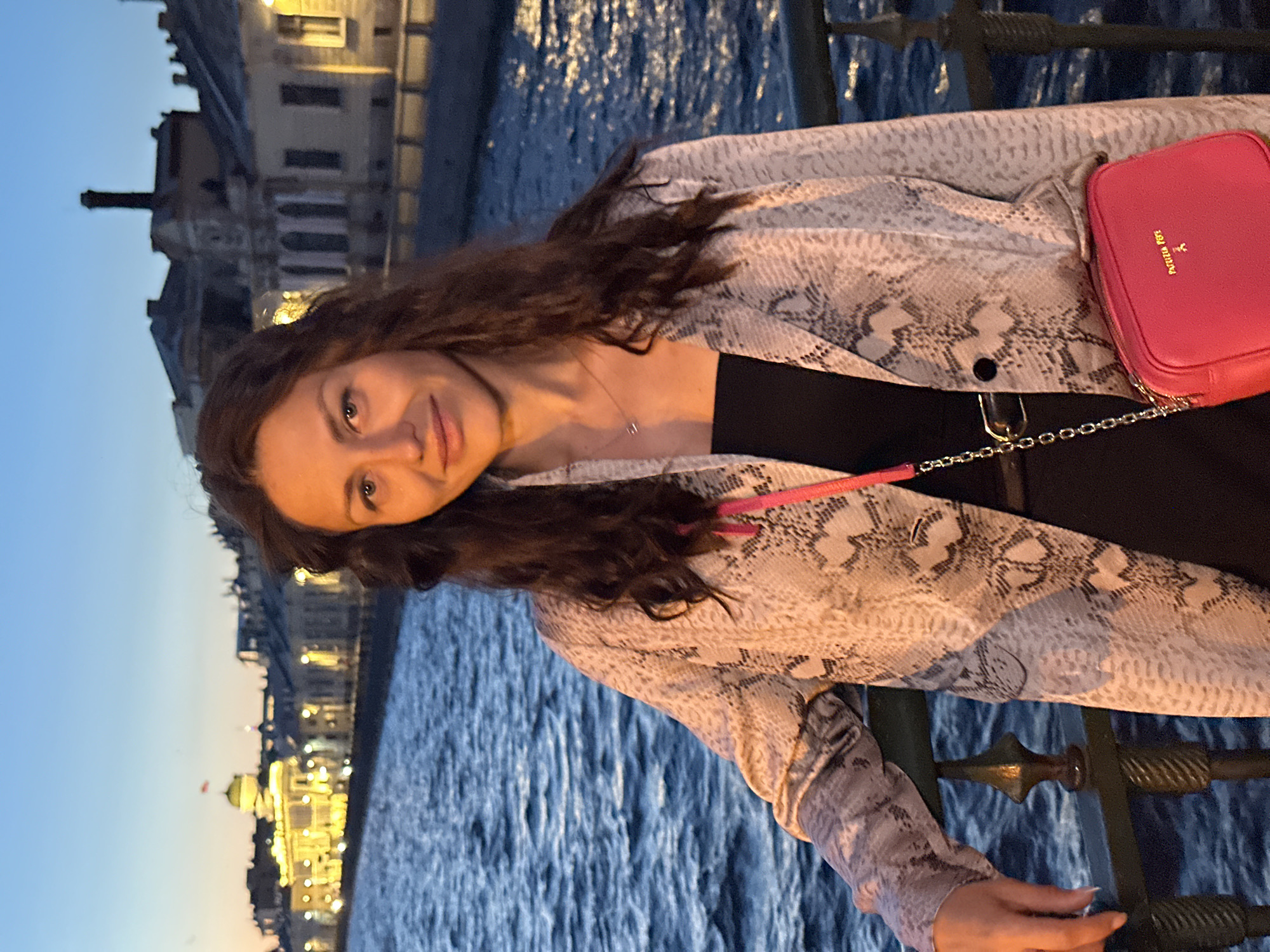 A medium shot shows a woman standing on a bridge at dusk in Saint Petersburg, Russia. She is leaning on the railing with her left arm. The woman has long, wavy brown hair and is wearing a snake-print blazer over a black strapless top, and a bright pink crossbody bag. The background shows a body of water, likely a canal or river, with buildings illuminated along the embankment. The sky is a gradient of blue and orange, indicating sunset or twilight. The buildings appear to be historical structures with a neoclassical architectural style. The water is rippled, suggesting a slight breeze.