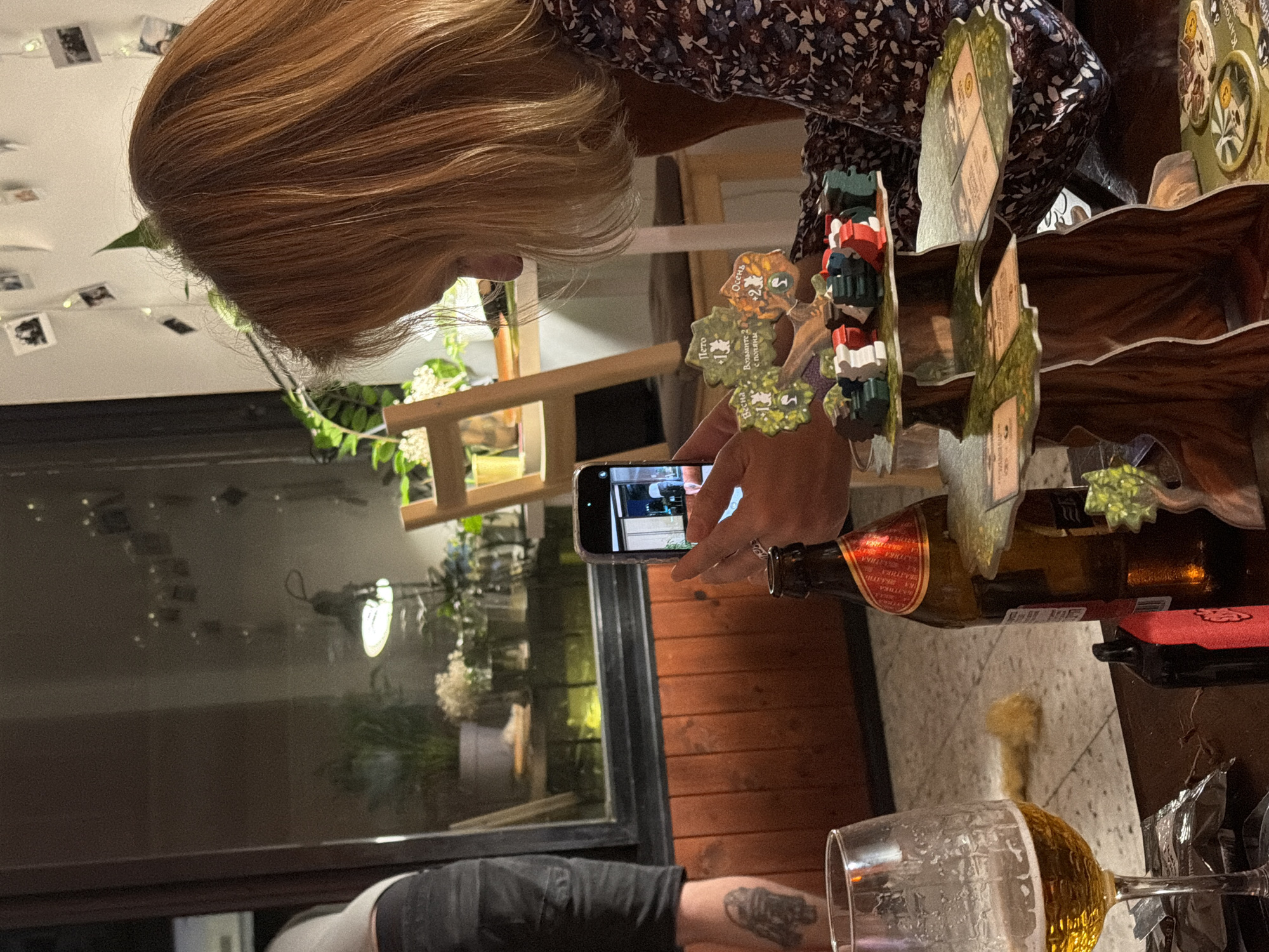 A woman with light brown hair is holding a smartphone and taking a photo of a wooden board game with several game pieces. The woman is wearing a dark, floral-patterned shirt. A beer bottle and a partially-filled wine glass are visible on the table. A person wearing dark pants and a light-colored shirt is visible in the bottom left. The scene appears to be indoors, near a window with a view of what appears to be a city at night. A trellis with plants is also visible behind the woman. Based on the visual clues, the location could be in Ramat Gan, Israel, but it is difficult to confirm definitively.