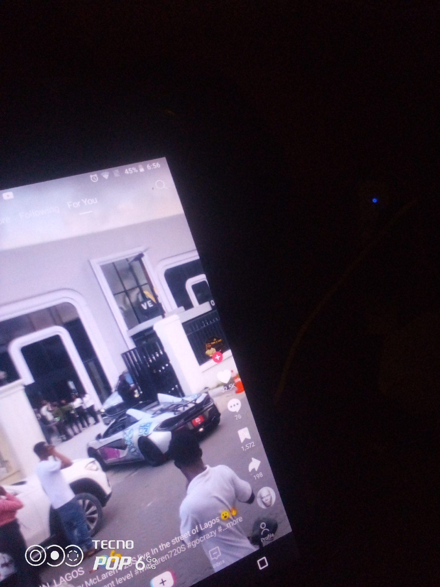 The image is a screenshot from a mobile phone displaying a social media application, likely TikTok, which shows a street scene. A white car is parked on the left, with a person in a white t-shirt and blue jeans standing next to it. In the background, a silver McLaren sports car with blue accents is parked. Several people are visible around the cars. The phone screen also shows a time of 6:56, battery at 45%, and indicators for "Following" and "For You." The text overlay on the bottom of the screen includes "IN LAGOS," "TECNO POP 6," "live In the street of Lagos," "by McLaren720s #gocrazy #...more," and "4G." The scene appears to be outdoors, with a building facade and white pillars visible. The overall lighting suggests it might be daytime, though the phone screen is illuminated. There is no specific information to place the location within the city of Howayi, Nigeria, as the text explicitly mentions Lagos.