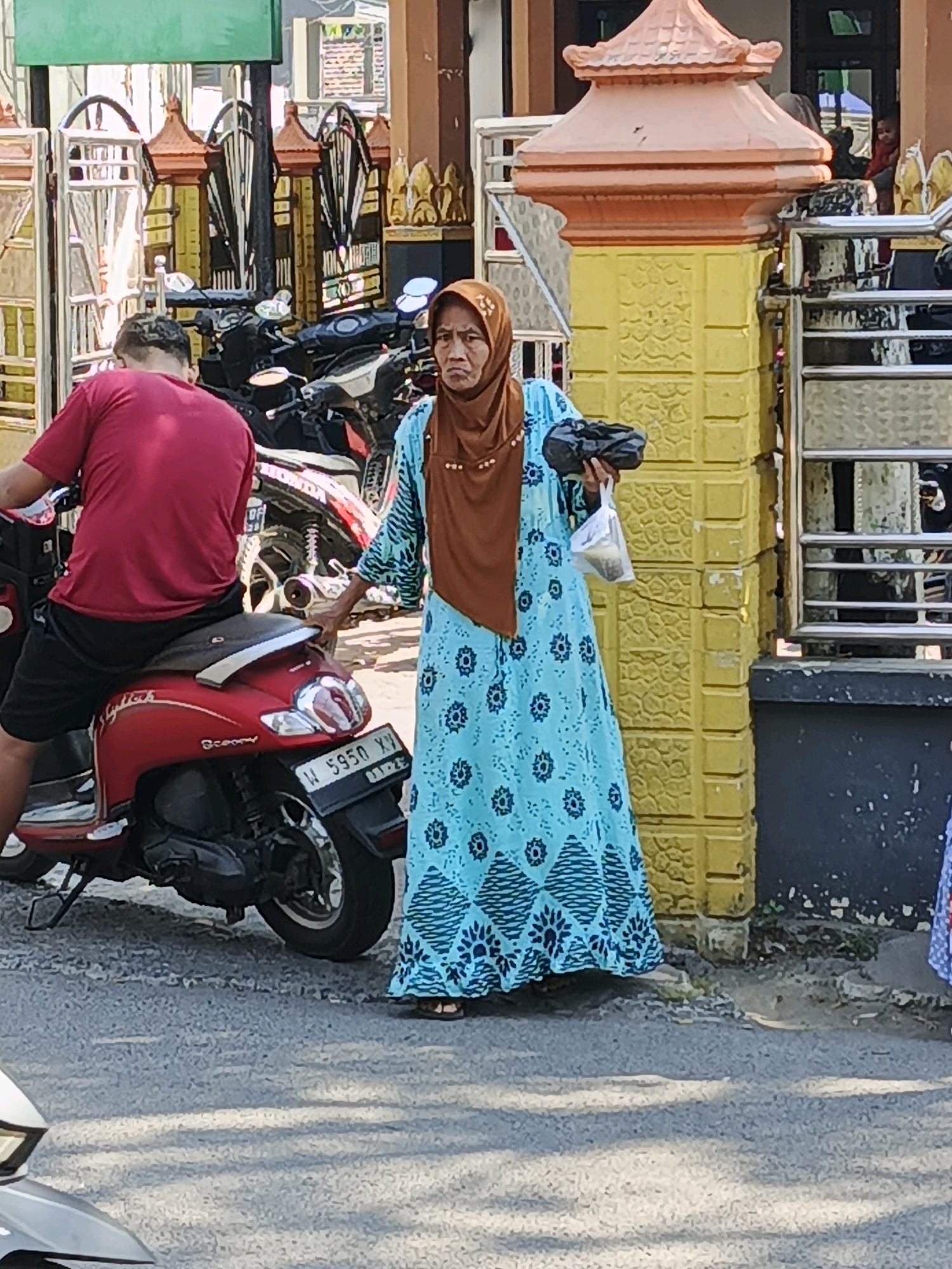 In Ketegan, Indonesia, a woman wearing a brown hijab and a blue patterned dress stands next to a red scooter. She holds a dark object in her left hand and a plastic bag in her right. A man wearing a red shirt and black shorts is seated on the scooter, facing away from the camera. The scooter has a license plate that reads "W 5950 XY". In the background, a yellow wall with textured patterns and a metal fence are visible. Several other motorcycles are parked behind the scooter. The ground is paved with asphalt and gravel.