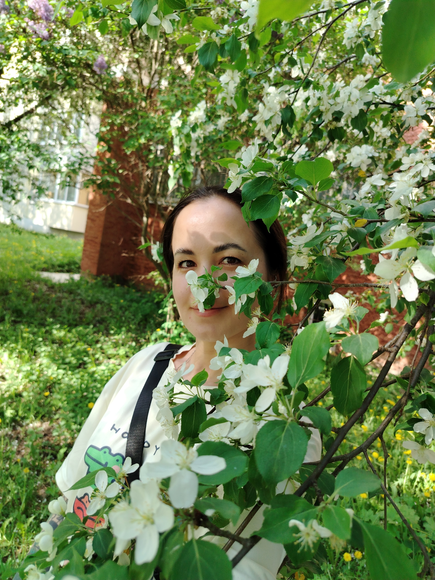 A woman stands among blooming branches, her face partially obscured by white blossoms and green leaves. She wears a light-colored t-shirt with a cartoon graphic and a black strap across her shoulder. The setting appears to be a park or garden, with green grass and flowering trees visible in the background. A brick building is partially visible behind the trees. The image was likely taken in Noviy, Russia.