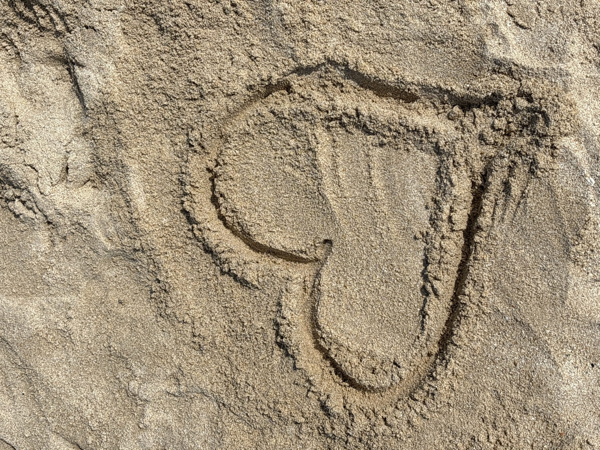 The image is a close-up, overhead shot of a heart shape drawn in the sand. The sand is a light brown color and appears to be somewhat damp, with visible grains and subtle ripples. The heart shape is outlined by a slightly deeper groove in the sand, creating a shadow that defines its form. The lighting suggests it is daytime, with sunlight casting shadows. There are no people or other objects visible in the frame. The image is focused entirely on the drawn heart in the sand. The location is not discernible from the image itself.