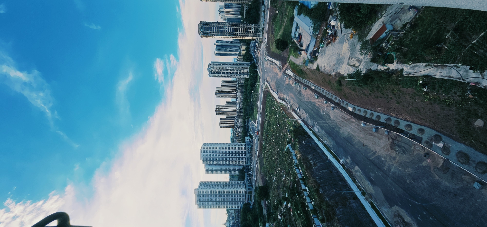 An aerial, vertical shot captures a partially developed urban landscape under a bright blue sky with scattered white clouds. In the background, a cluster of tall, modern residential buildings stands prominently. In the mid-ground, a wide, paved road winds through the scene, appearing to be under construction or recently completed, with concrete barriers and lighting poles lining its edges. The immediate foreground and the areas adjacent to the road are characterized by uneven terrain, patches of grass, and visible construction debris, suggesting ongoing development. A few vehicles are sparsely distributed on the road. The overall impression is that of a growing urban area with infrastructure development in progress. The location is identified as Jiulongpo District, China.