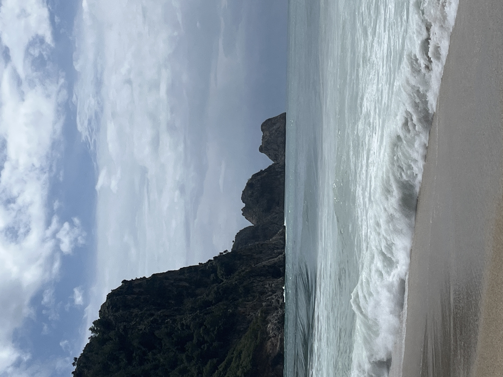 The image shows a beach scene in Alanya, Turkey. A dark, vegetation-covered mountain rises from the left side of the frame, leading to rocky cliffs. The right side of the image depicts a beach with white waves crashing onto the sand. The sky above shows a mix of blue sky and white clouds.