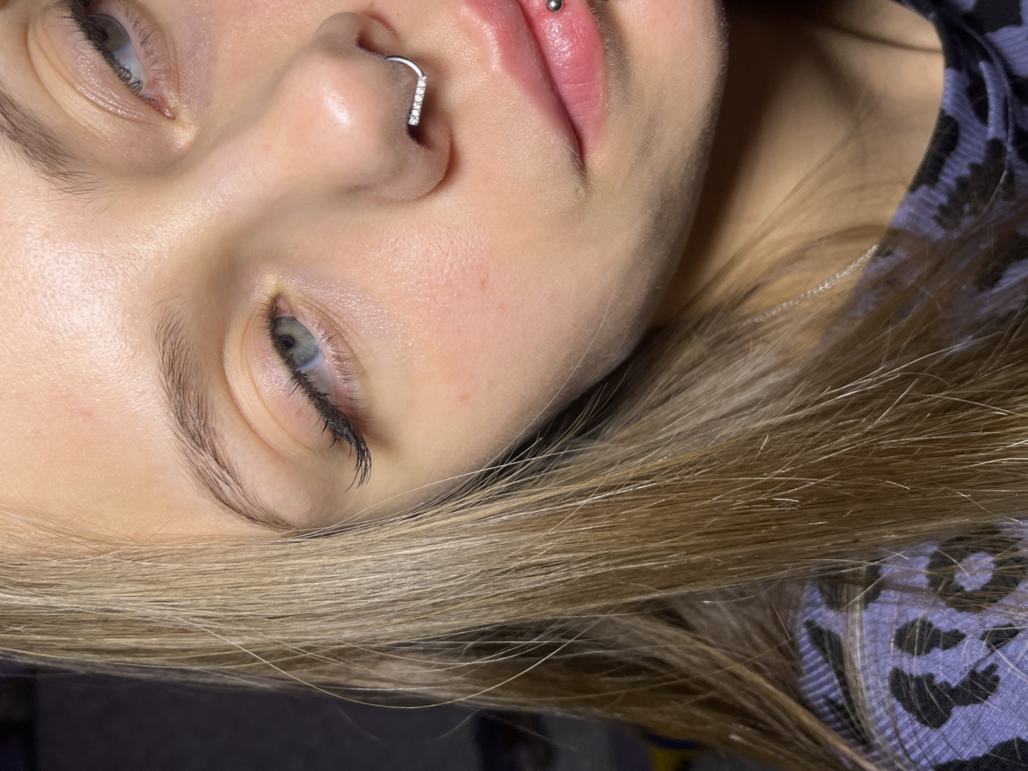 A close-up, high-angle portrait shows a young woman with fair skin and light blonde hair. Her face is tilted toward the upper left corner. She has piercings in her nose and upper lip. The focus is on her face, with details of her skin texture, eye color, and piercings clearly visible. She is wearing a patterned shirt or top. The background is out of focus, with only hints of color and texture visible. There is no information to suggest that this image was taken in Фоки, Russia.