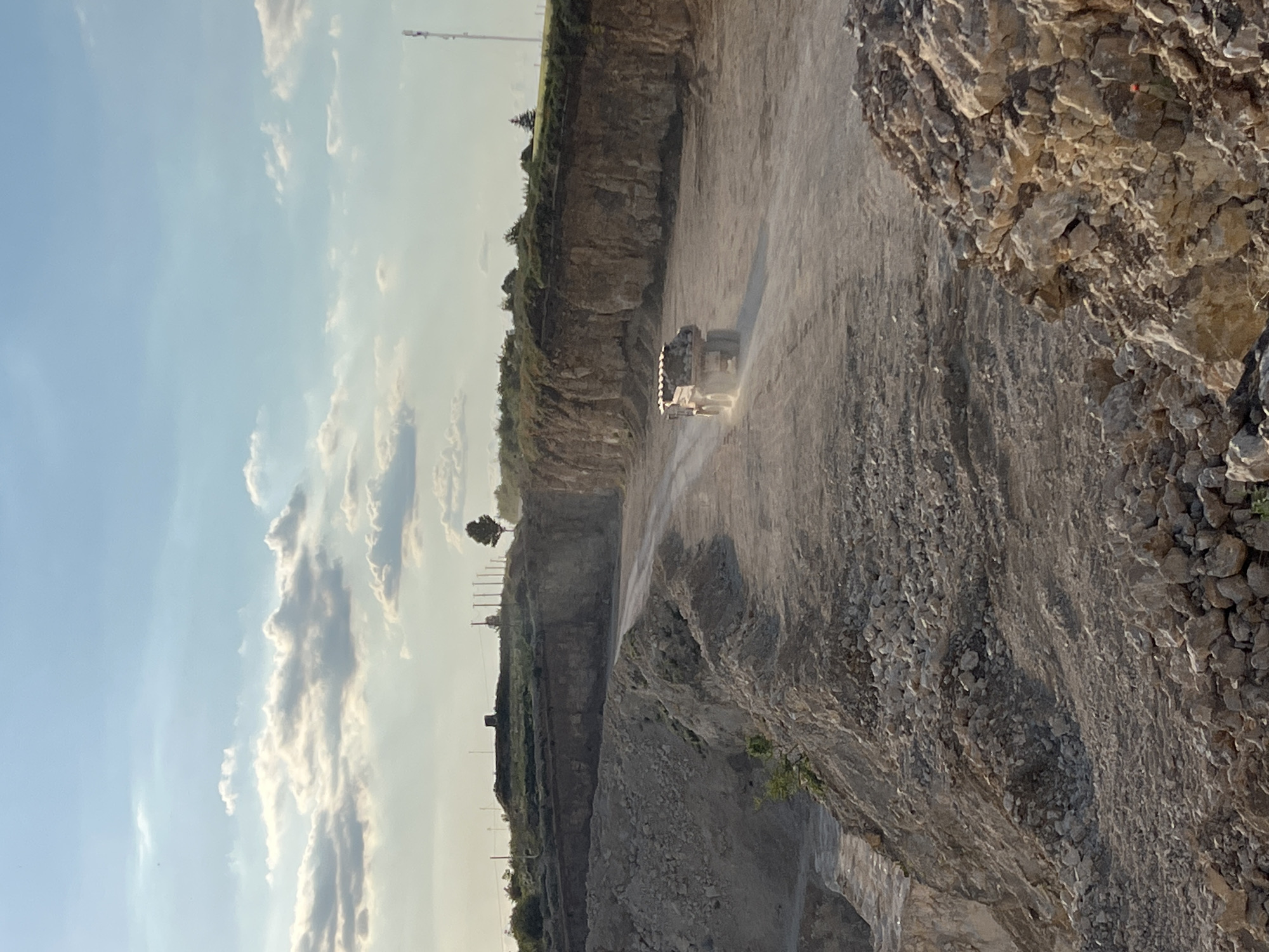The image depicts a quarry with a large dump truck driving on a dirt road. The truck is loaded with rocks, and dust is being kicked up by its tires. The quarry walls are steep and rocky, with vegetation growing on the upper edge. The sky is blue with some clouds, and the sun appears to be low in the sky, suggesting it is either early morning or late afternoon. The scene is consistent with an active mining or construction site. The specific location of Khomivka, Ukraine is not discernible from the image itself.