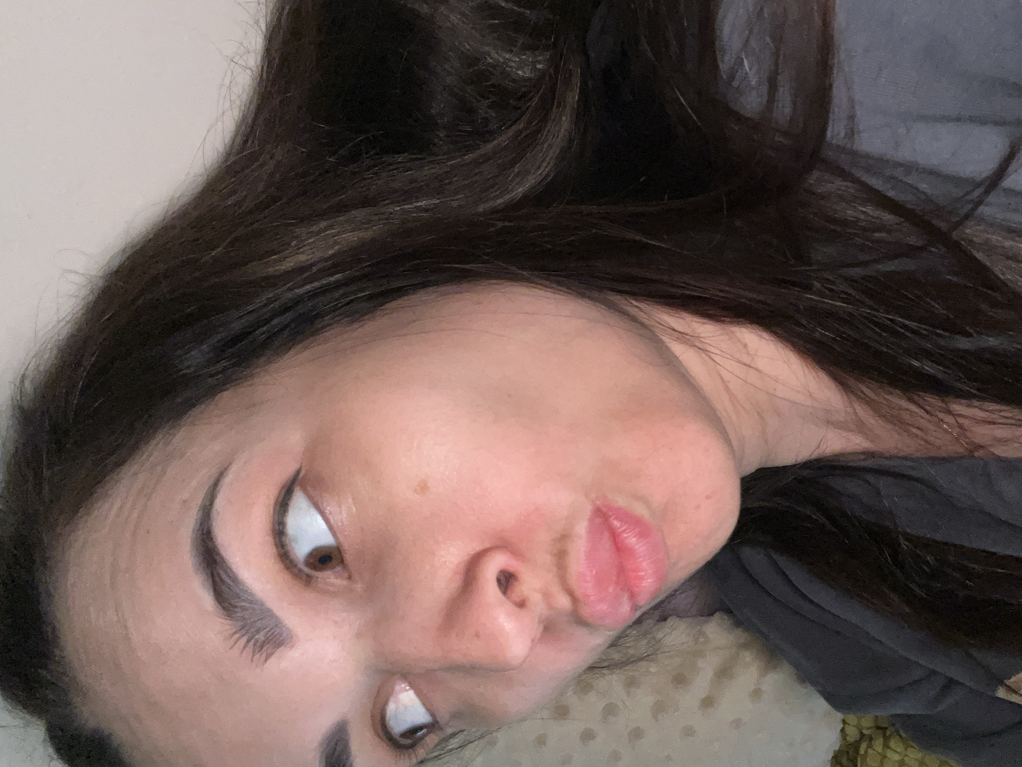 A medium close-up shot focuses on the face of a woman with long, dark brown hair. Her head is tilted, and she is looking off to the left with her eyes wide open. Her lips are pursed in a "kiss face" or "duck face" expression. A mole is visible on her right cheek. She is wearing a dark gray shirt. The background is softly lit and appears to be a bed with a patterned pillow. The image is taken from a slightly high angle.