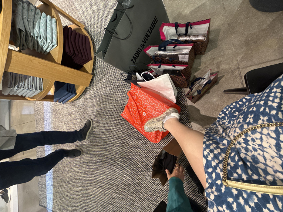 A cluttered floor with shopping bags from Zadig & Voltaire and other stores. A rotating display rack with folded clothing stands to the left. Two people are partially visible: one with dark jeans and shoes, and another wearing a blue patterned dress and sparkly sneakers, holding a shoe box. The floor is covered with a neutral-toned rug.