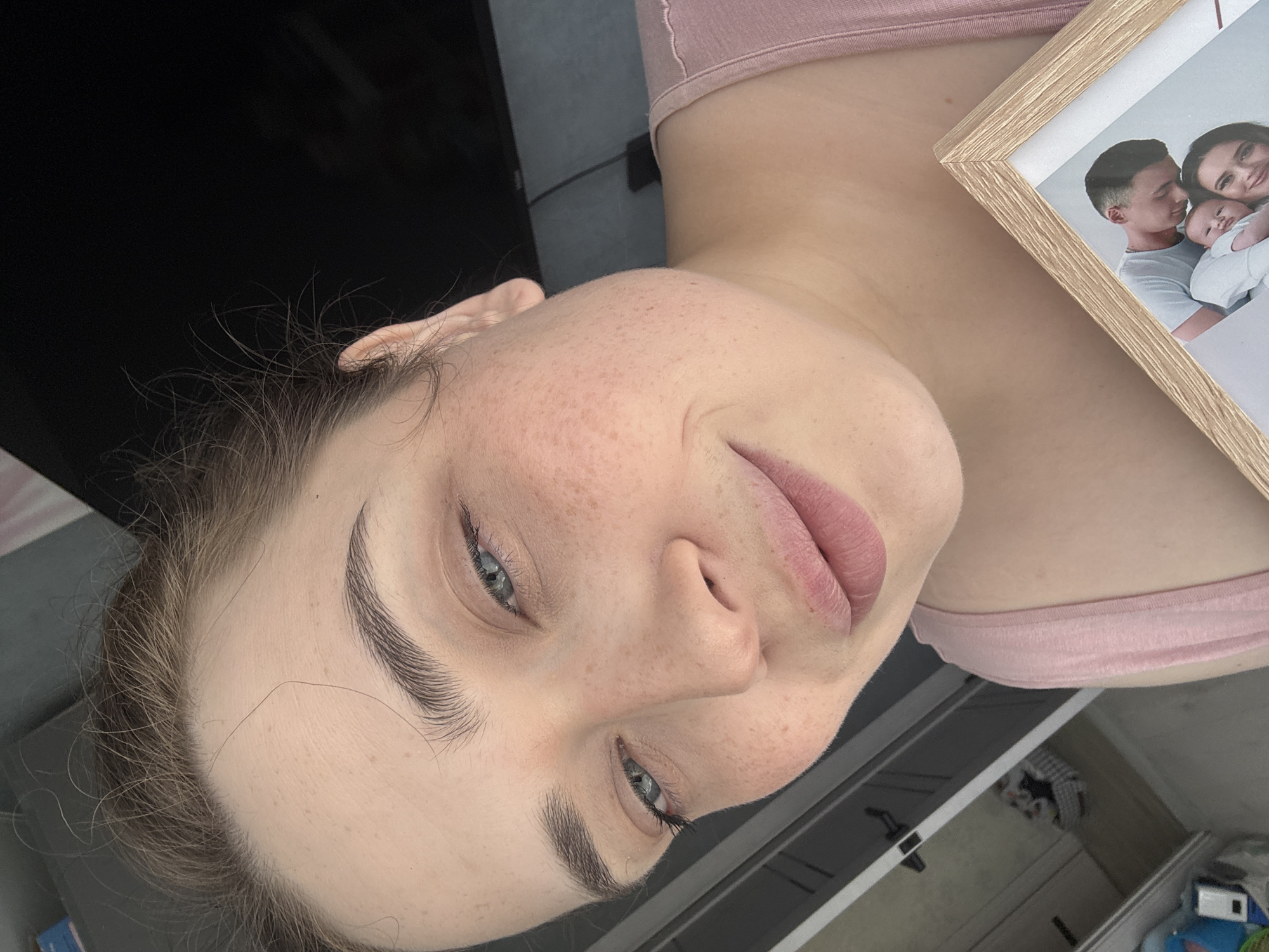 A close-up, slightly tilted portrait of a woman with fair skin, freckles, and light blue eyes. She has well-defined eyebrows and her lips are light pink. She is wearing a light pink top. A framed photograph containing a family is on the right side of the image. The background includes a dark TV screen, a wall, and a glimpse of a room with a possible closet. The location is unknown and may or may not be Chaikovsky, Russia.