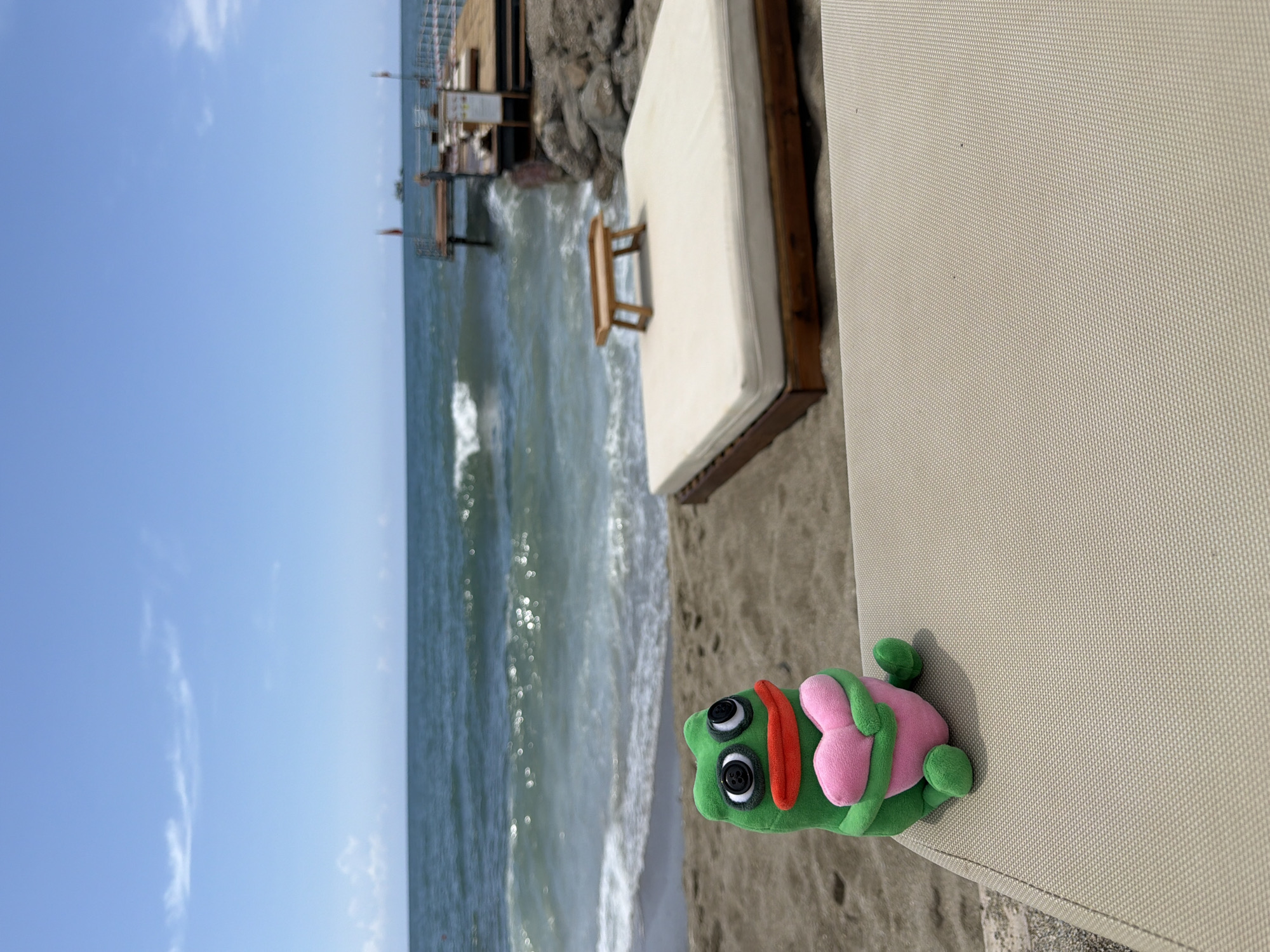 A sunny daytime shot depicts a stuffed green frog toy with pink appendages lying on a light-colored woven surface, likely a lounge chair, with a beach and ocean view. The beach has light-colored sand. In the background, there is a weathered wooden pier or structure extending into the water. The sky is blue with scattered clouds. The location is likely Alanya, Turkey, as implied by the prompt.