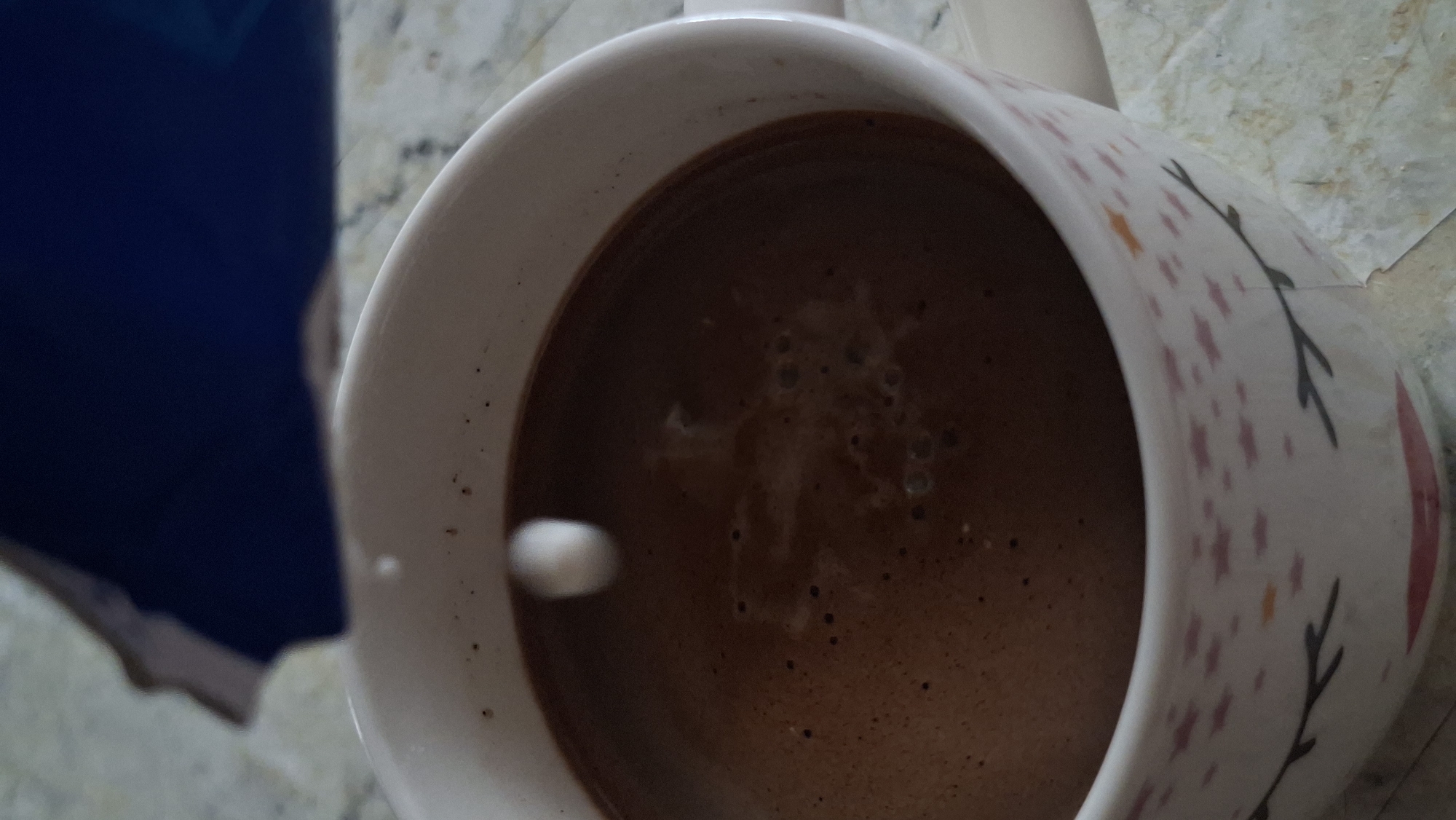 A close-up, overhead shot captures a mug of dark, frothy liquid, likely coffee or hot chocolate, on a speckled countertop. A single drop of white liquid, possibly milk or cream, is suspended in mid-air just above the surface of the drink, creating a ripple effect. The mug itself is white with a pattern of pink stars and a simple face design on its lower half, featuring closed eyes and lips. In the upper portion of the image, a dark blue object, possibly a book or container, is partially visible. The lighting suggests an indoor setting, likely a kitchen or dining area, within the city of Забалоцкі сельскі Савет, Belarus. No people are present in the frame.